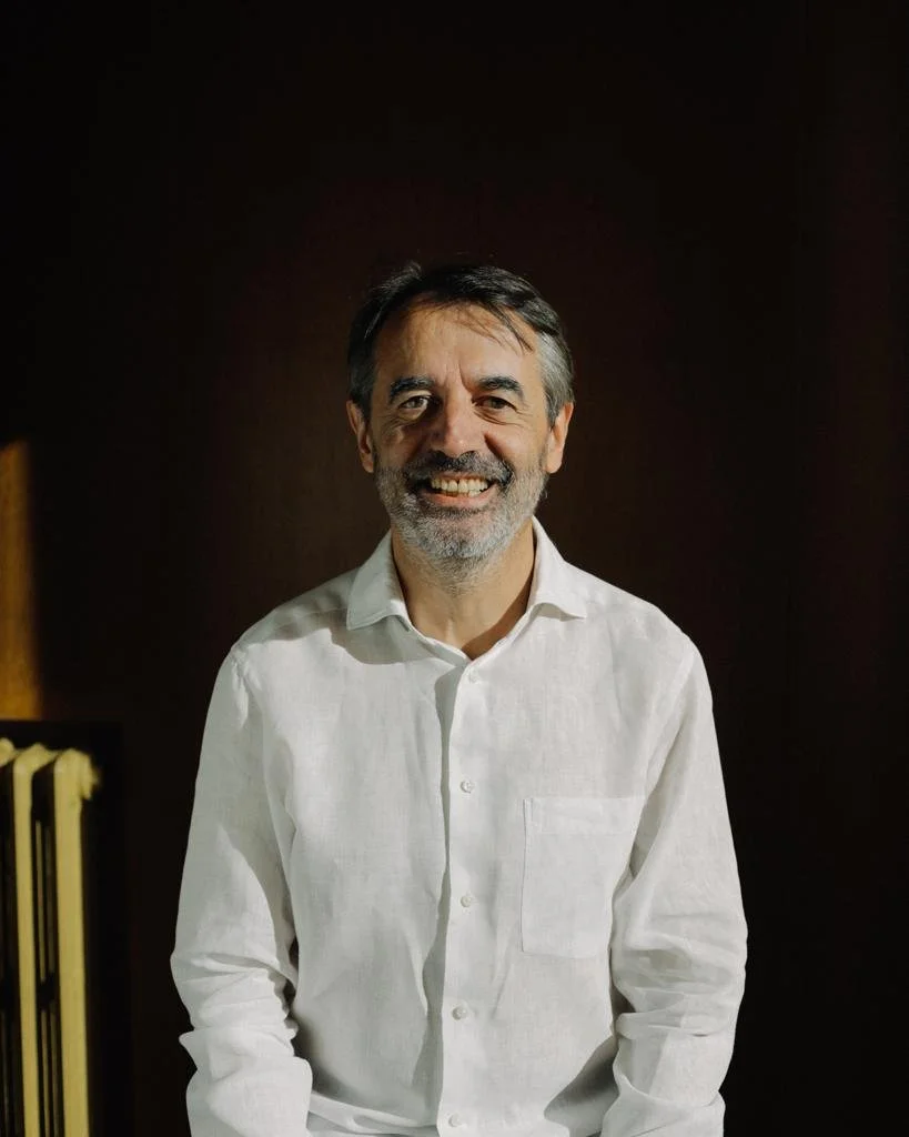 Hombre sonriendo con barba y pelo gris, usando camisa blanca, fondo oscuro.