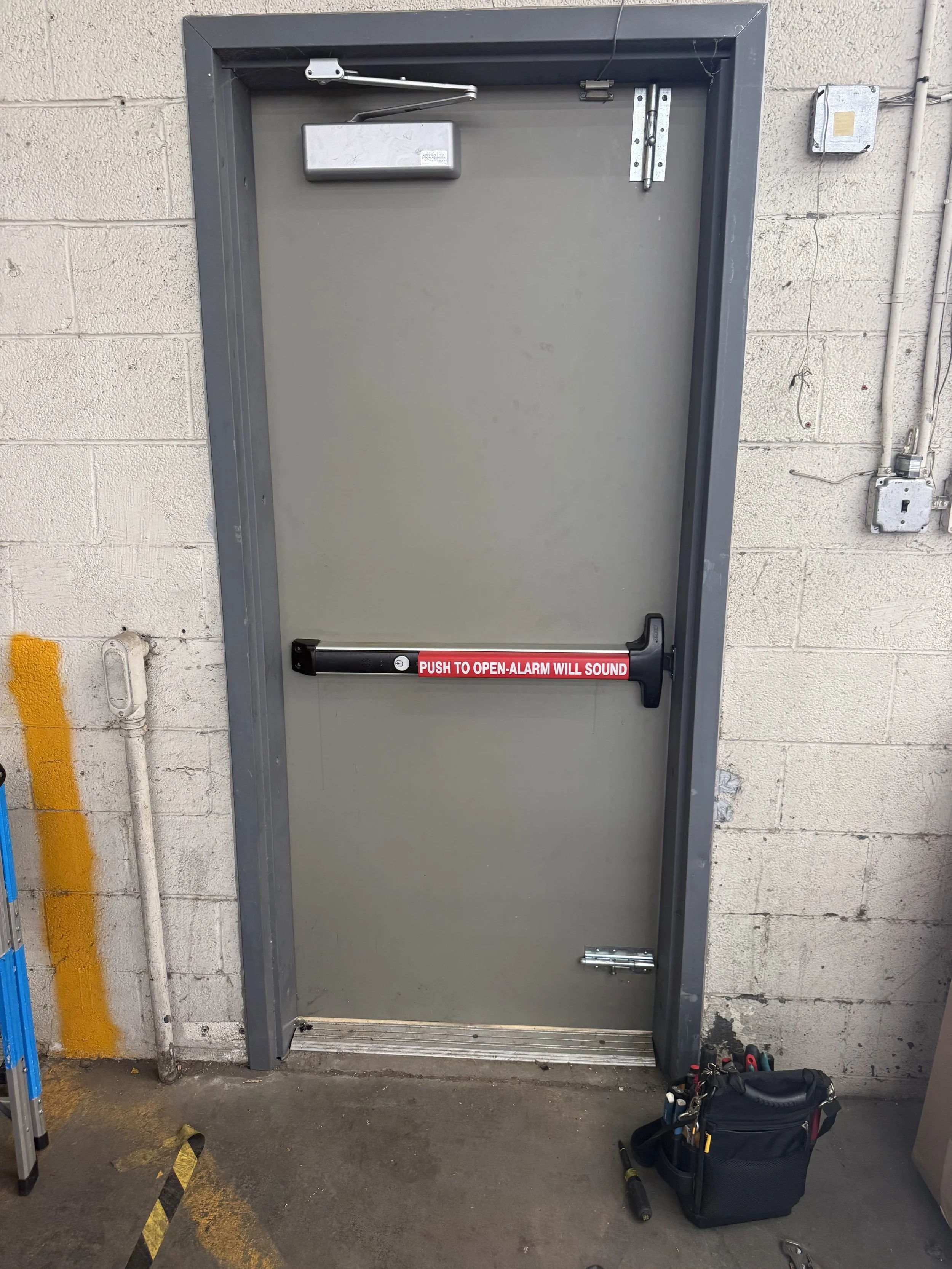 A gray emergency exit door with a push bar that has a red label reading 'PUSH TO OPEN-ALARM WILL SOUND' in a garage or industrial area. There is a black bag, screwdriver, and tools on the floor near the door.