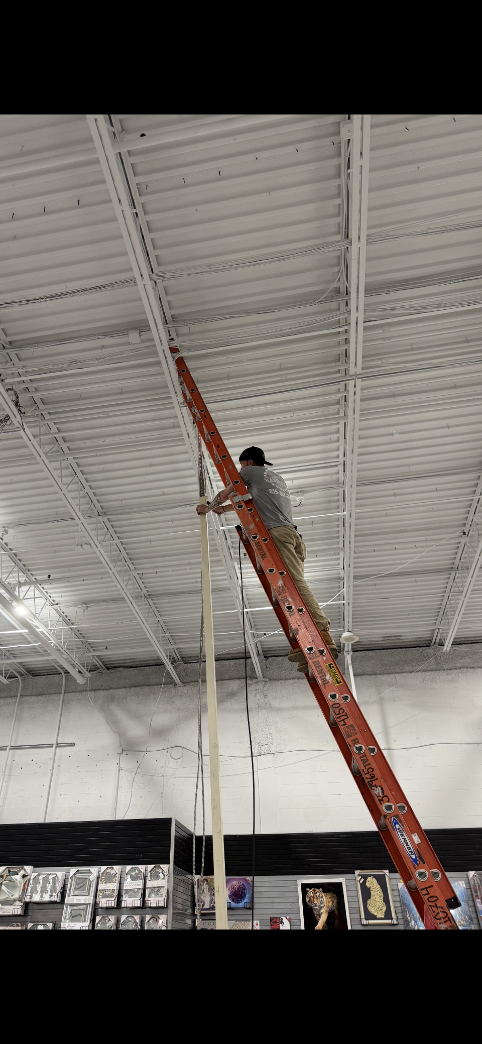 A man standing on a red extension ladder installing or repairing a ceiling in a retail store. The ceiling has exposed industrial-style metal beams and wiring, and shelves with decorative items and artwork are visible below.