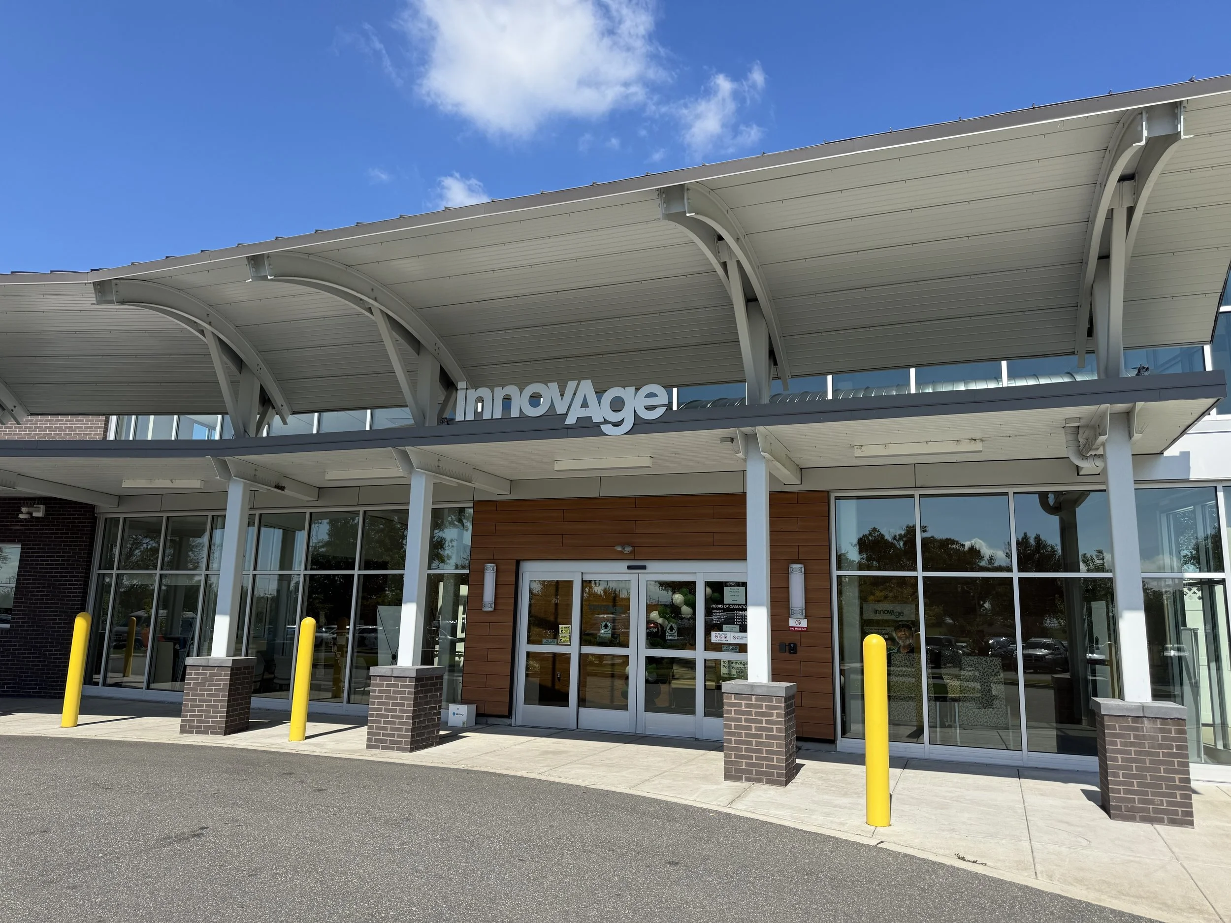 The exterior of a modern building with the sign 'innovaGe' on the upper part. The building has a curved white roof, glass doors, and brick pillars. The sky is partly cloudy.