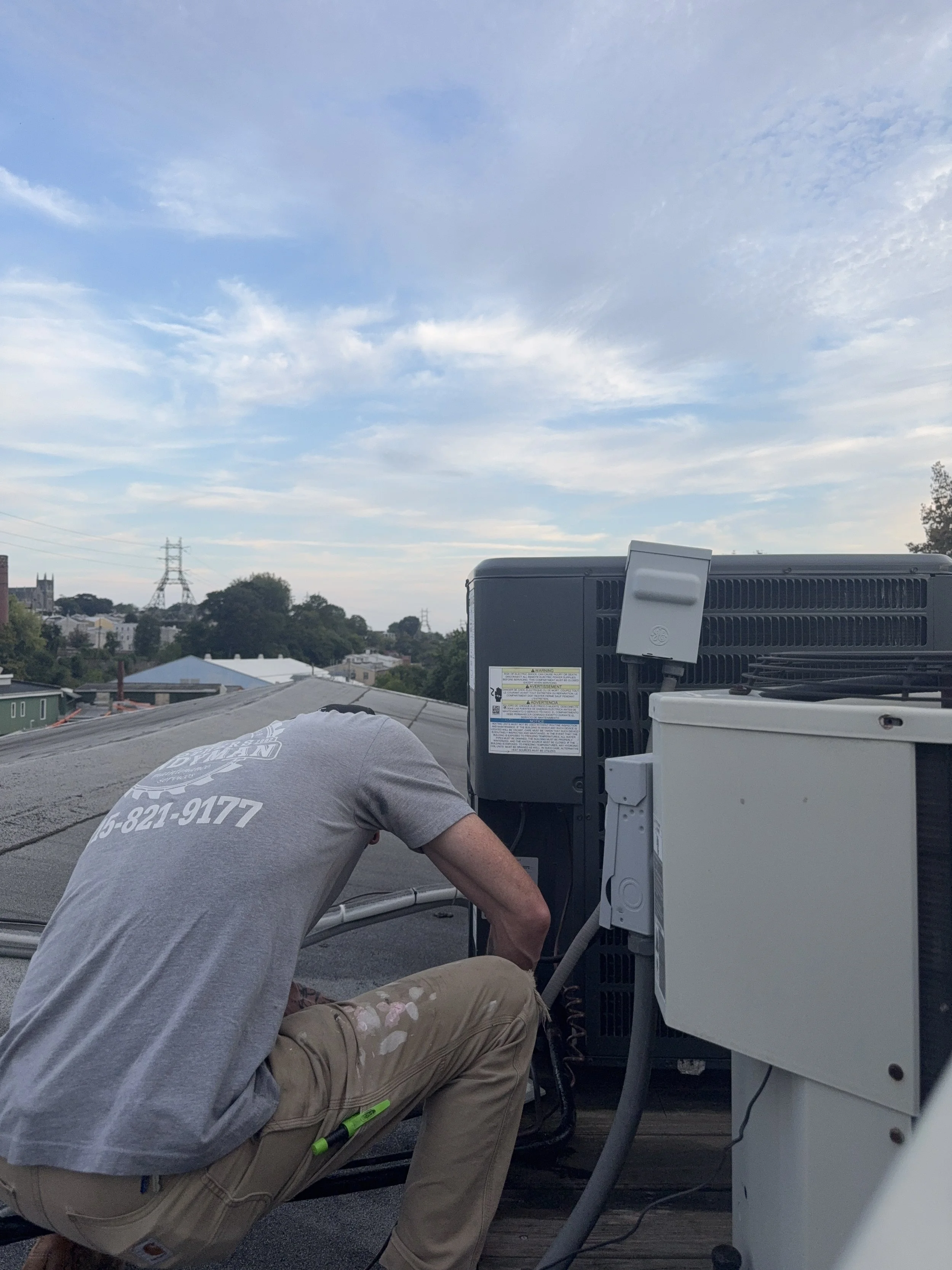 A worker wearing a gray shirt and beige pants kneels down on a rooftop repairing or maintaining HVAC or electrical equipment, with houses, trees, and power lines visible in the background under a partly cloudy sky.