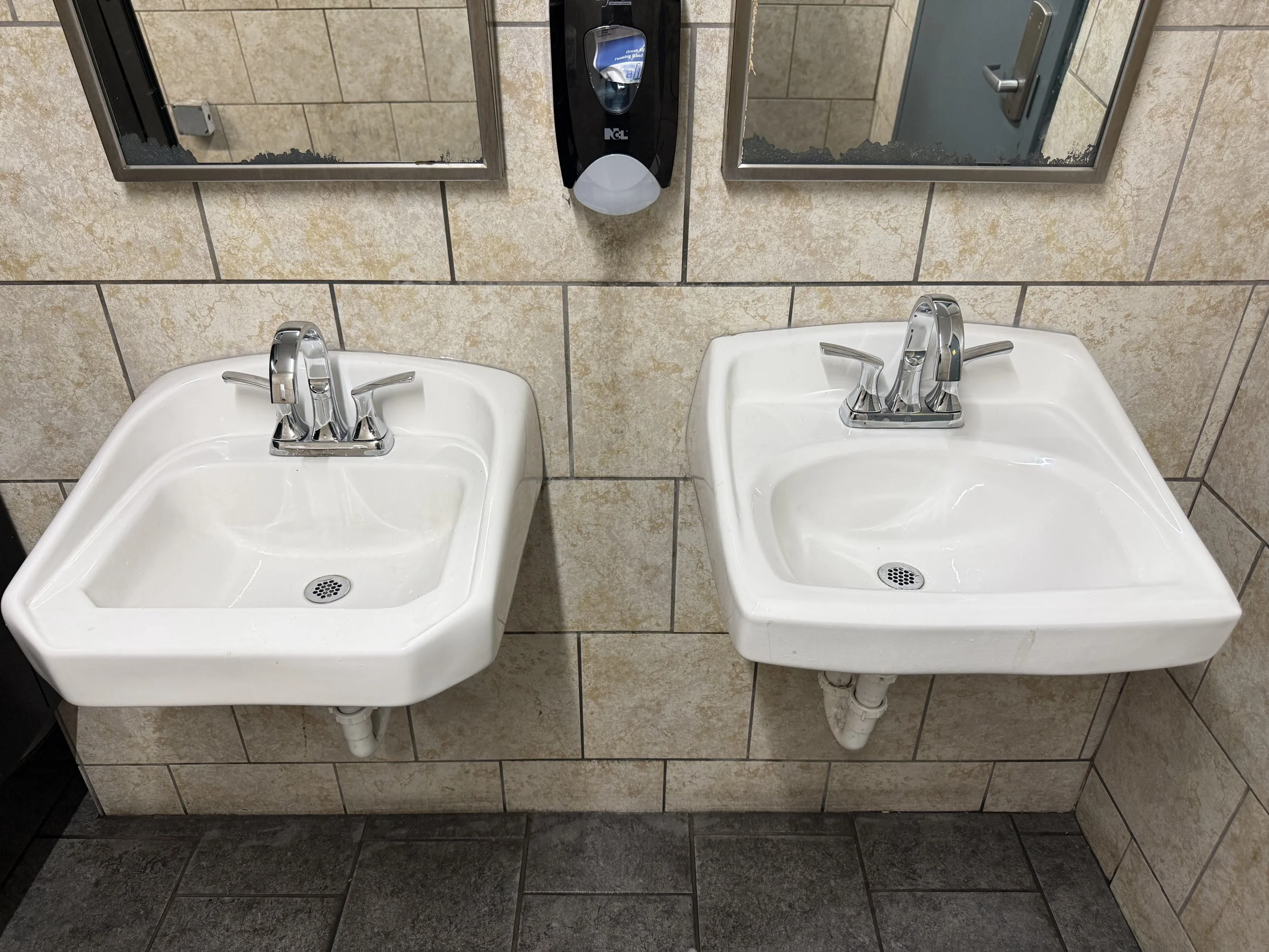 A bathroom with two white sinks side by side, each with a chrome faucet, mounted on a beige tiled wall. There are two mirrors above each sink and a black soap dispenser between them. The sinks are slightly dirty with water stains and debris at the drains, and the pipes underneath are exposed.