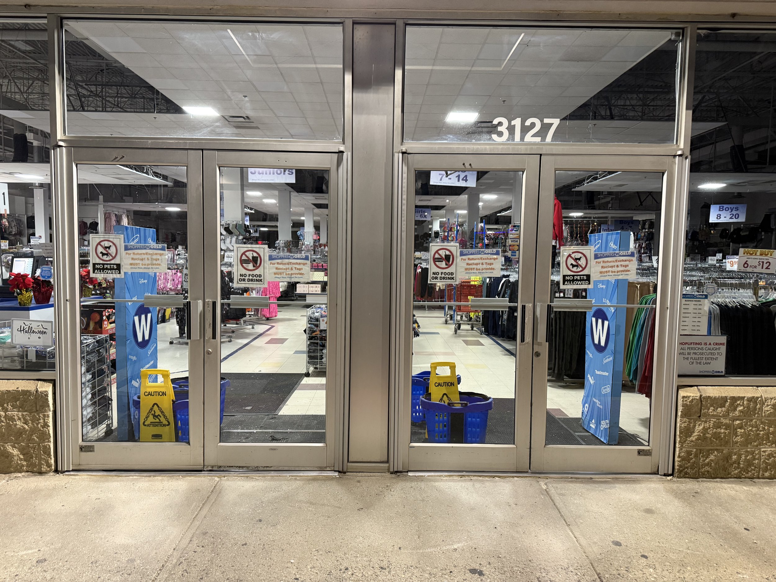 Store entrance with glass doors, inside are clothing racks, signs for boys and girls sections, and yellow caution signs on the floor.