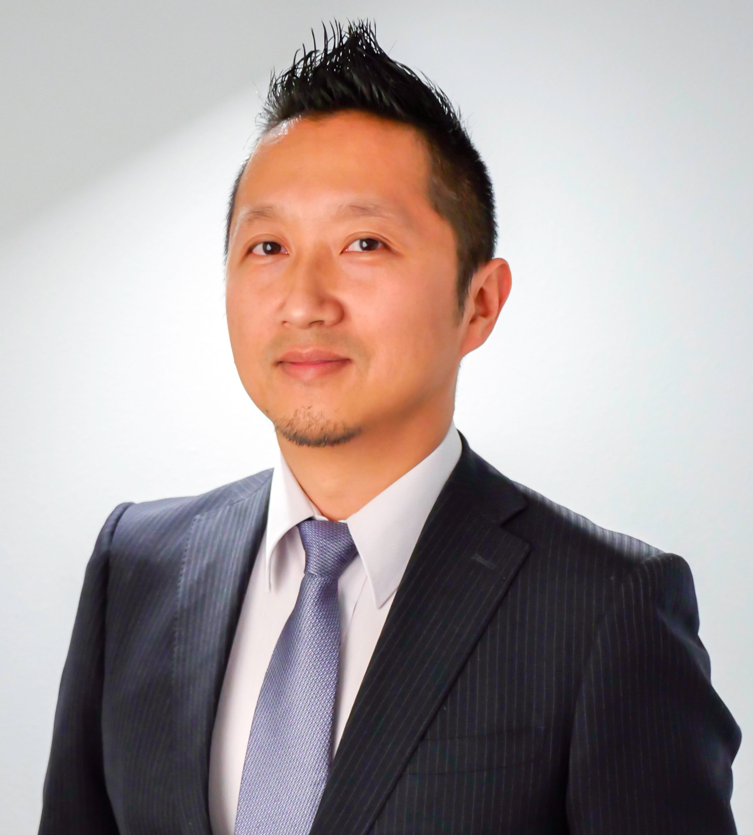 A man in a dark pinstripe suit with a purple tie and white shirt, posing against a plain light background.
