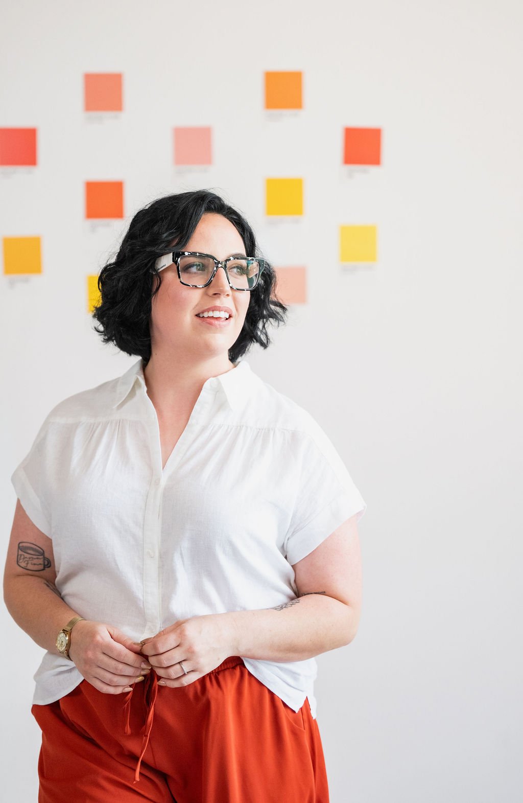 A woman with short black hair and glasses, wearing a white shirt and orange pants, standing in front of a wall with colorful sticky notes.