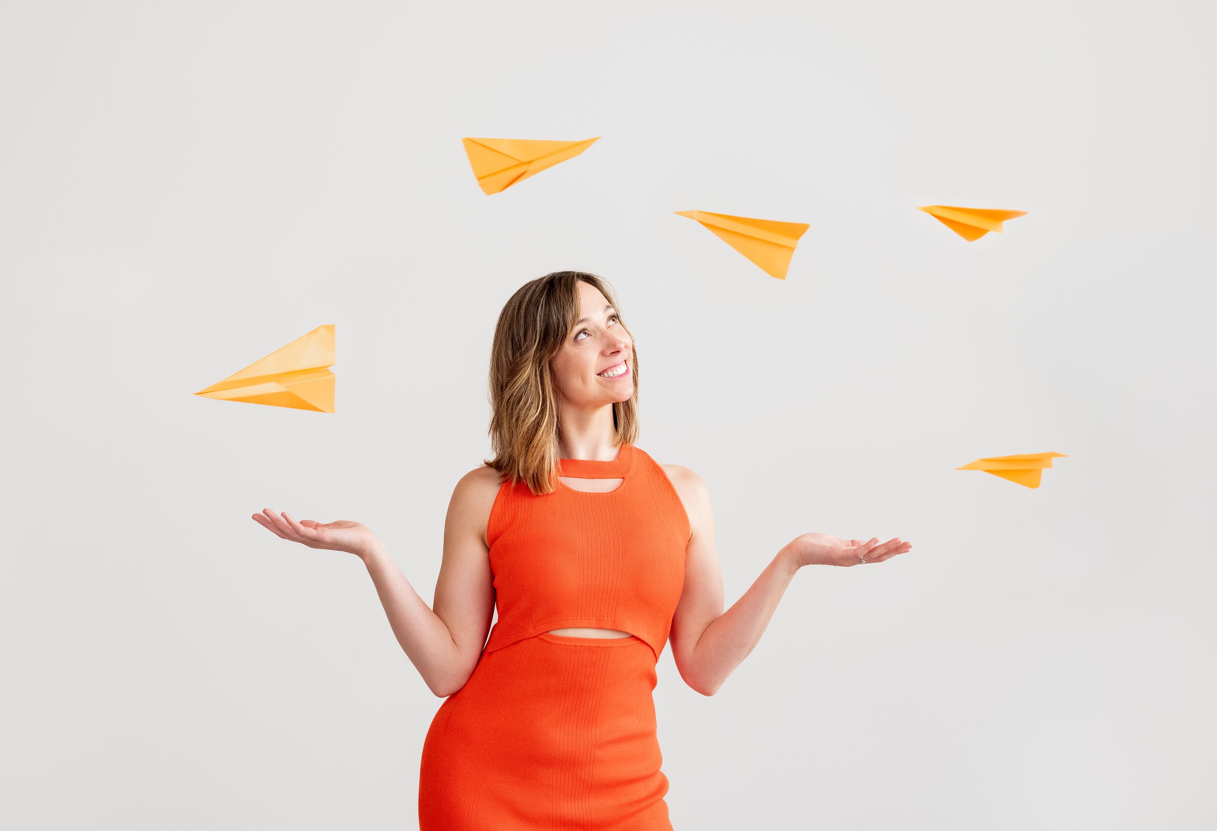 A woman with shoulder-length hair wearing an orange sleeveless dress, standing with her arms outstretched and a smile on her face, surrounded by orange paper planes floating in the air against a plain white background.