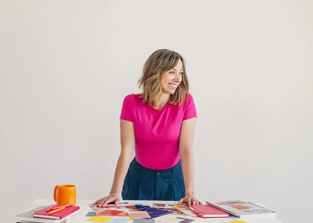 A woman wearing a pink shirt and blue trousers leaning on a white table with notebooks, papers, color samples, and an orange mug, smiling and looking to her left, against a plain white background.