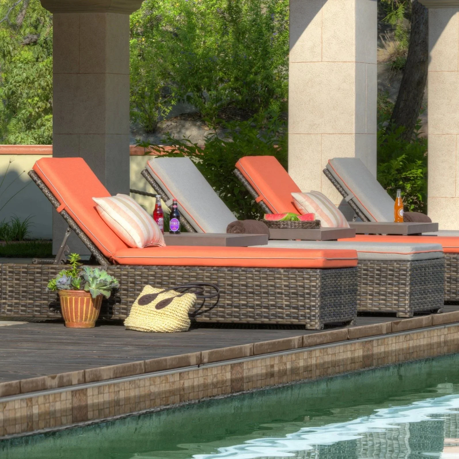 Poolside area with four lounge chairs, two with orange cushions and two with gray cushions, each with a colorful pillow. There are drinks on the tables next to some of the chairs and a potted plant on the deck. The pool with greenish water is visible