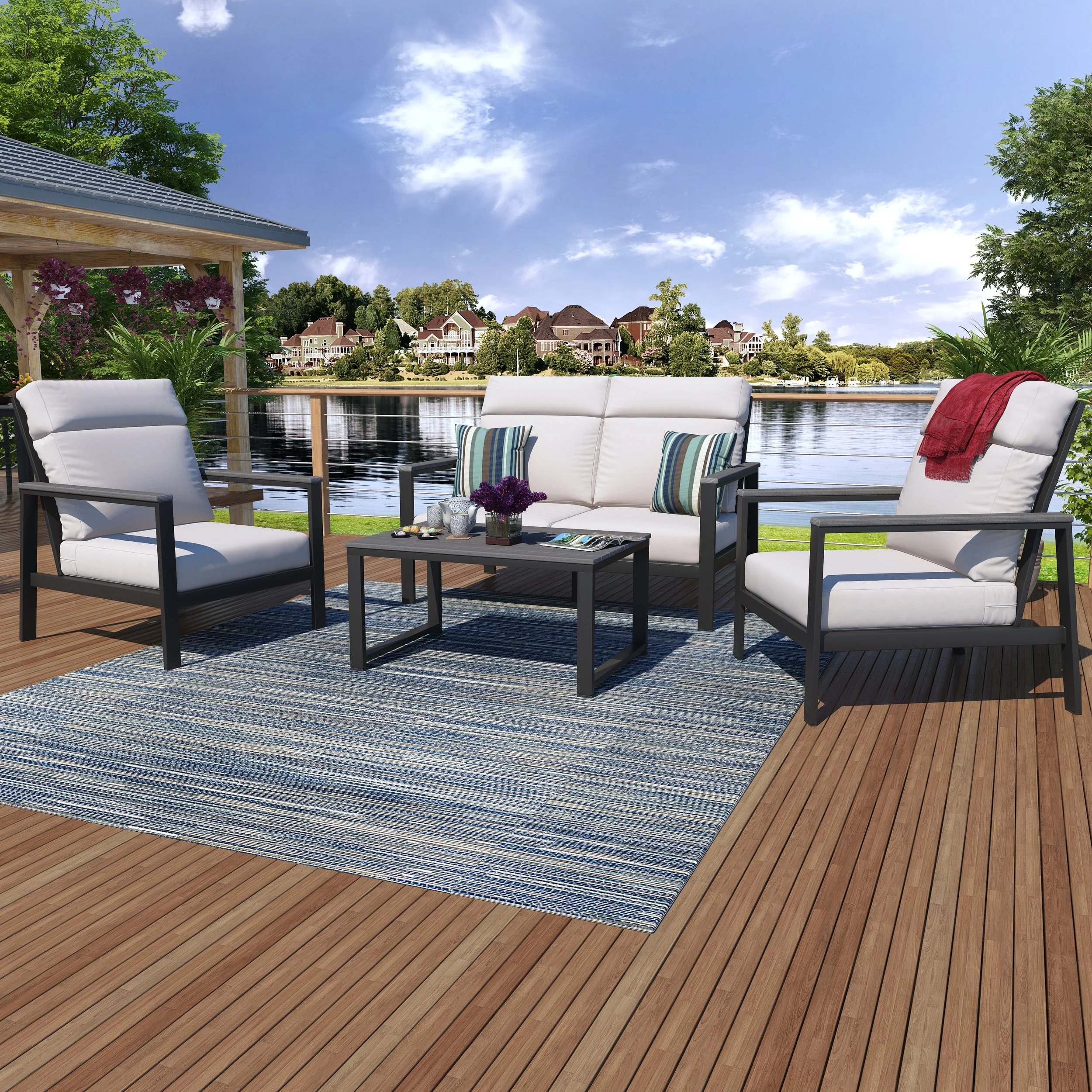 Image of an outdoor riverfront seating area with white cushioned chairs, a black coffee table with decorative vases, a blue and white striped rug, and greenery including trees and flowering plants, against a backdrop of houses across the water under 