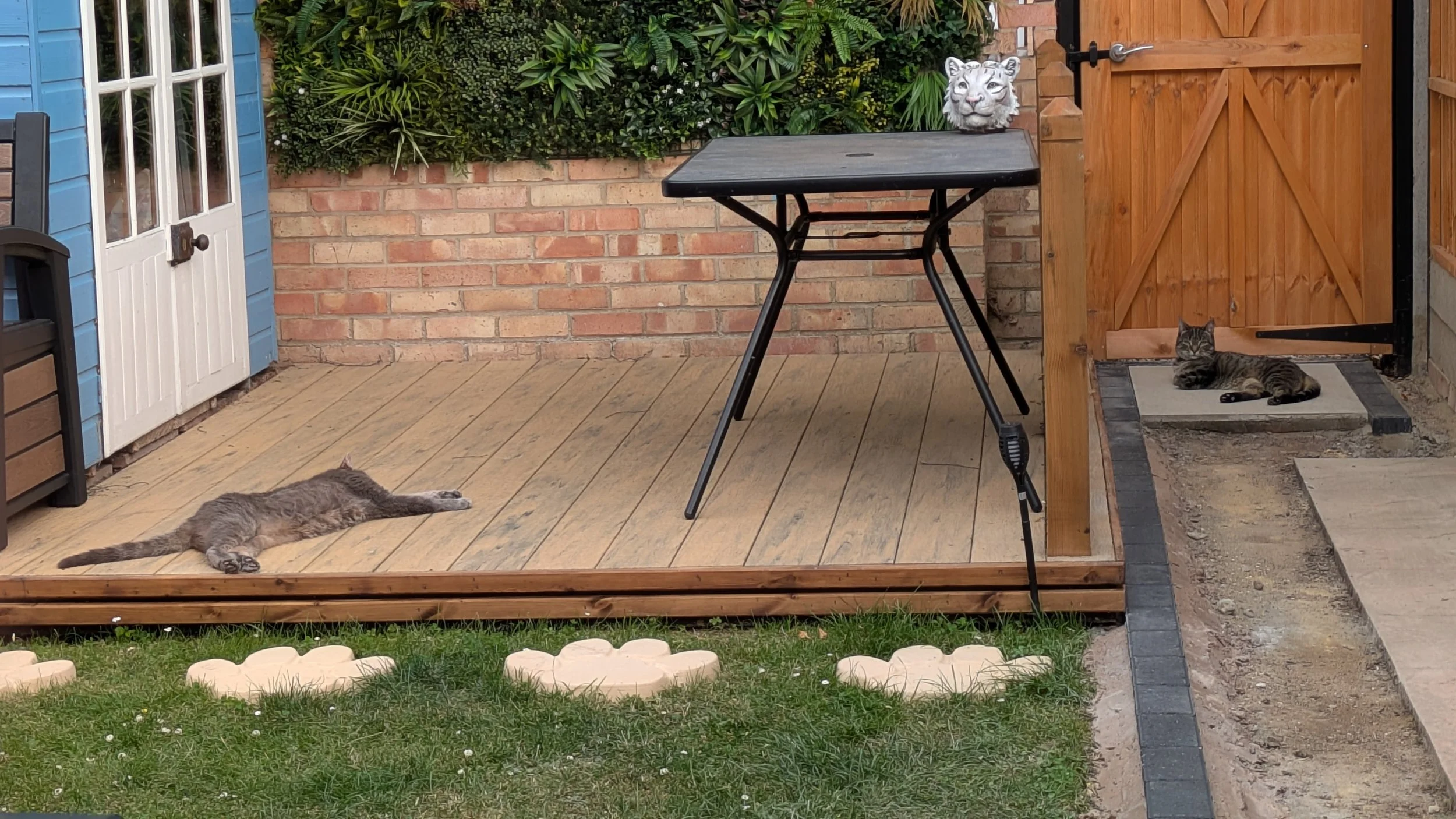 Composite decking with a brick wall, greenery, and a wooden fence and gate. Garden table, with porcelain tiled footpath