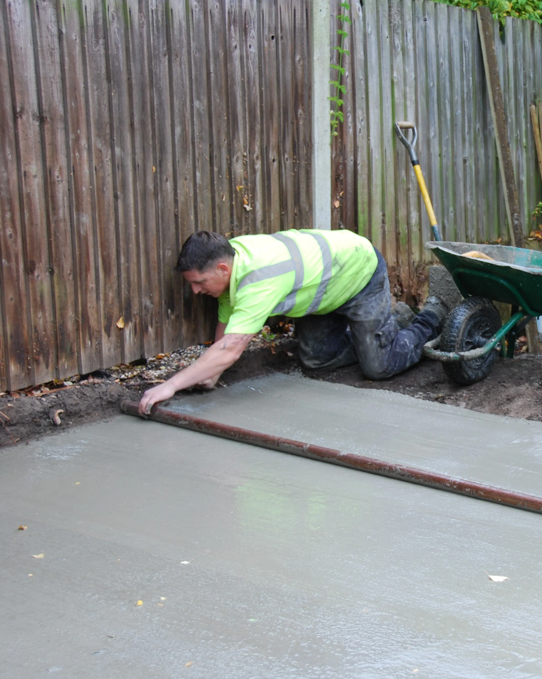 Concrete base, smoothing the surface with a trowel. The person is wearing a high-visibility yellow vest and dark pants. A wheelbarrow nearby contains concrete, and a shovel is propped against a wooden fence.