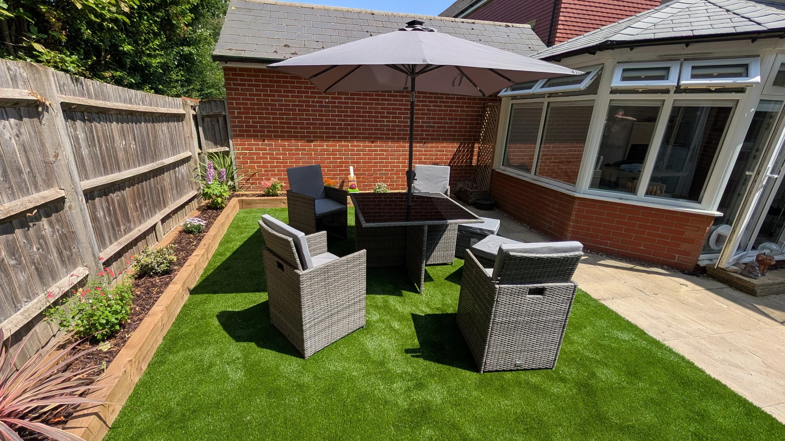 Backyard patio area with outdoor furniture, large umbrella, brick house, flower bed with plants, and a fenced yard.