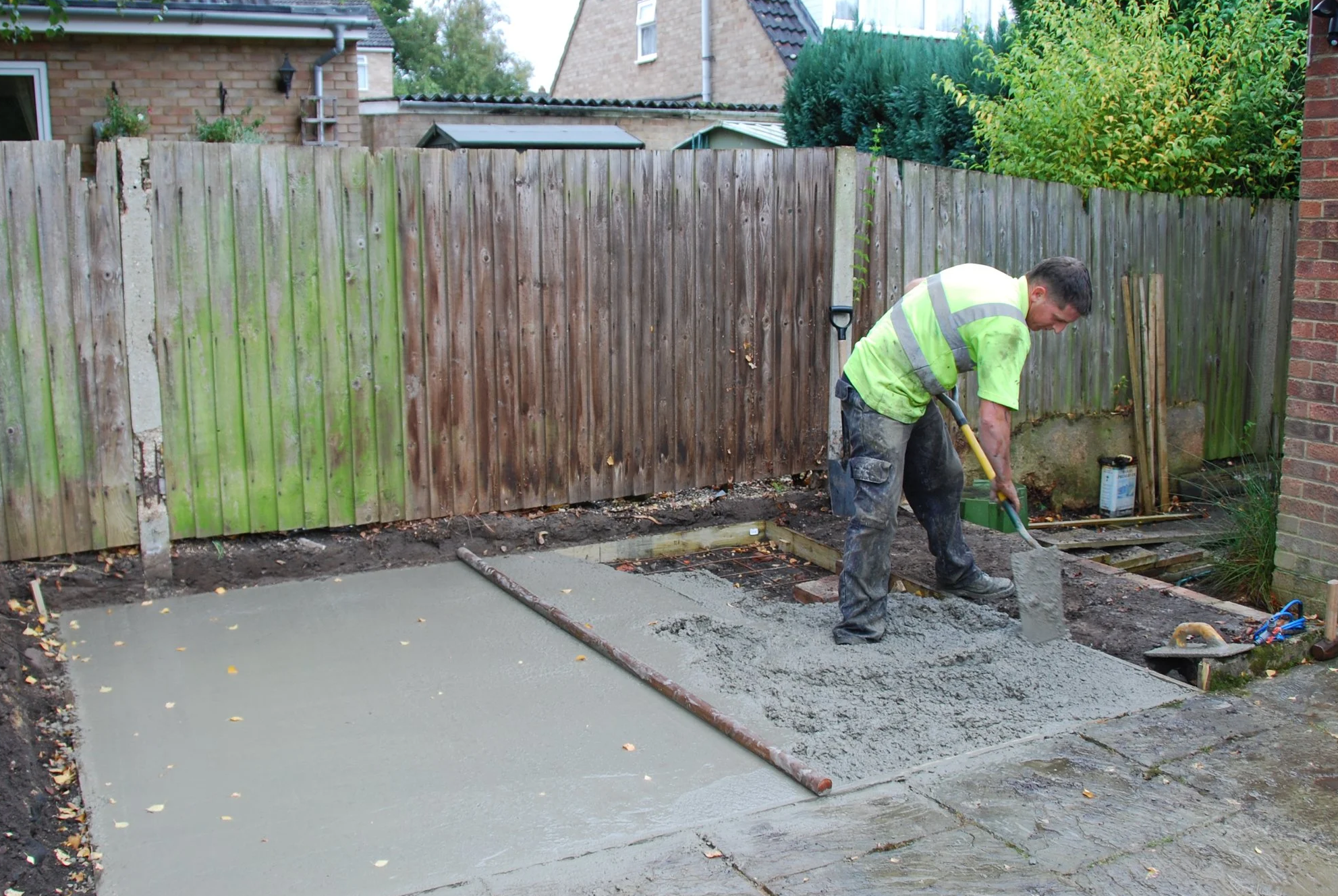Man working on a concrete patio in a backyard, with a wooden fence and houses in the background.