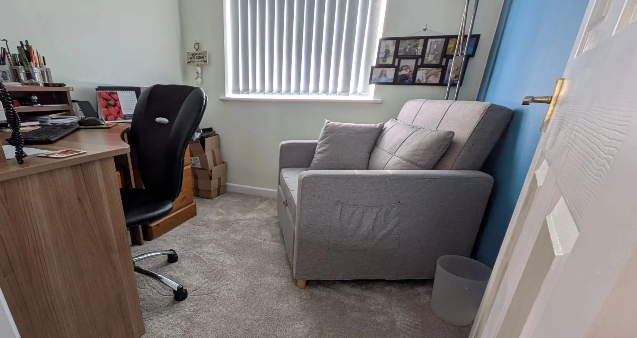 Home office with a desk, black office chair, grey seat, window with vertical blinds, and various office supplies.