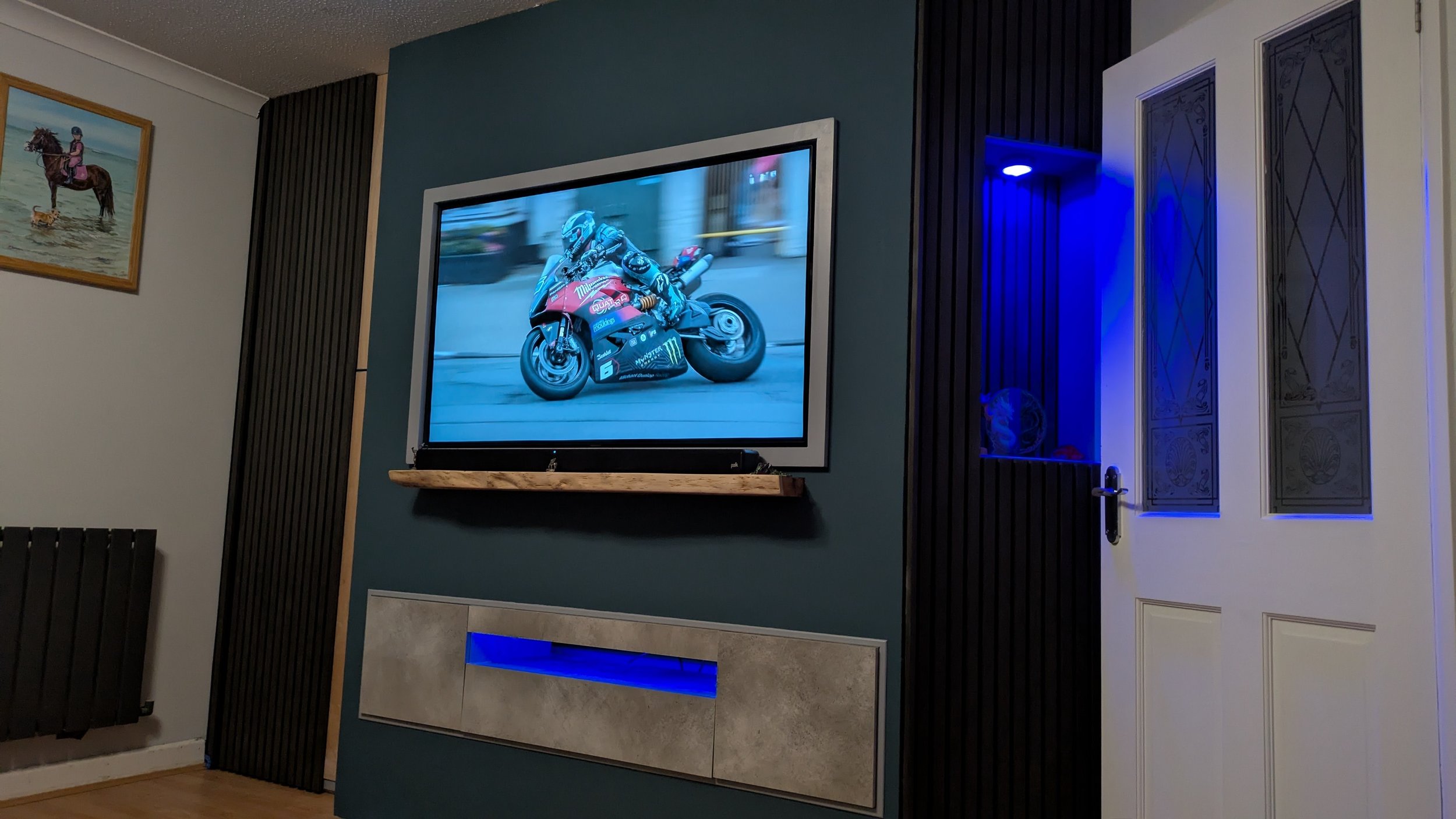 Media wall with a wall-mounted TV, acoustic sound panels, built in cupboard and shelving unit, illuminated by blue light.