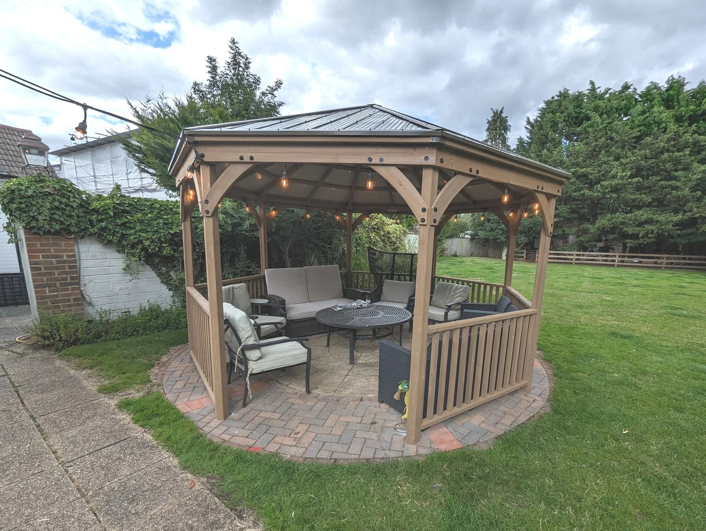 A backyard with a wooden gazebo featuring string lights, outdoor seating, potted plants, trees, and a brick and concrete patio on a cloudy day.