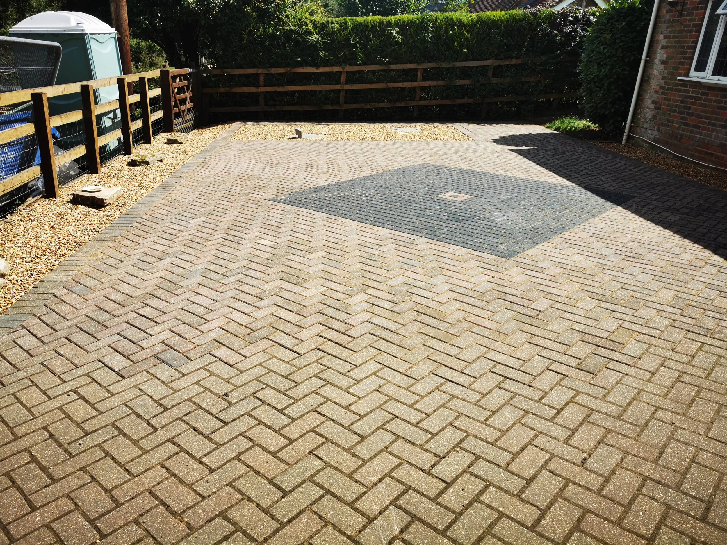 A paved outdoor area with brick and concrete pavers, a wooden fence, a lawn, and a brick house wall with a window.