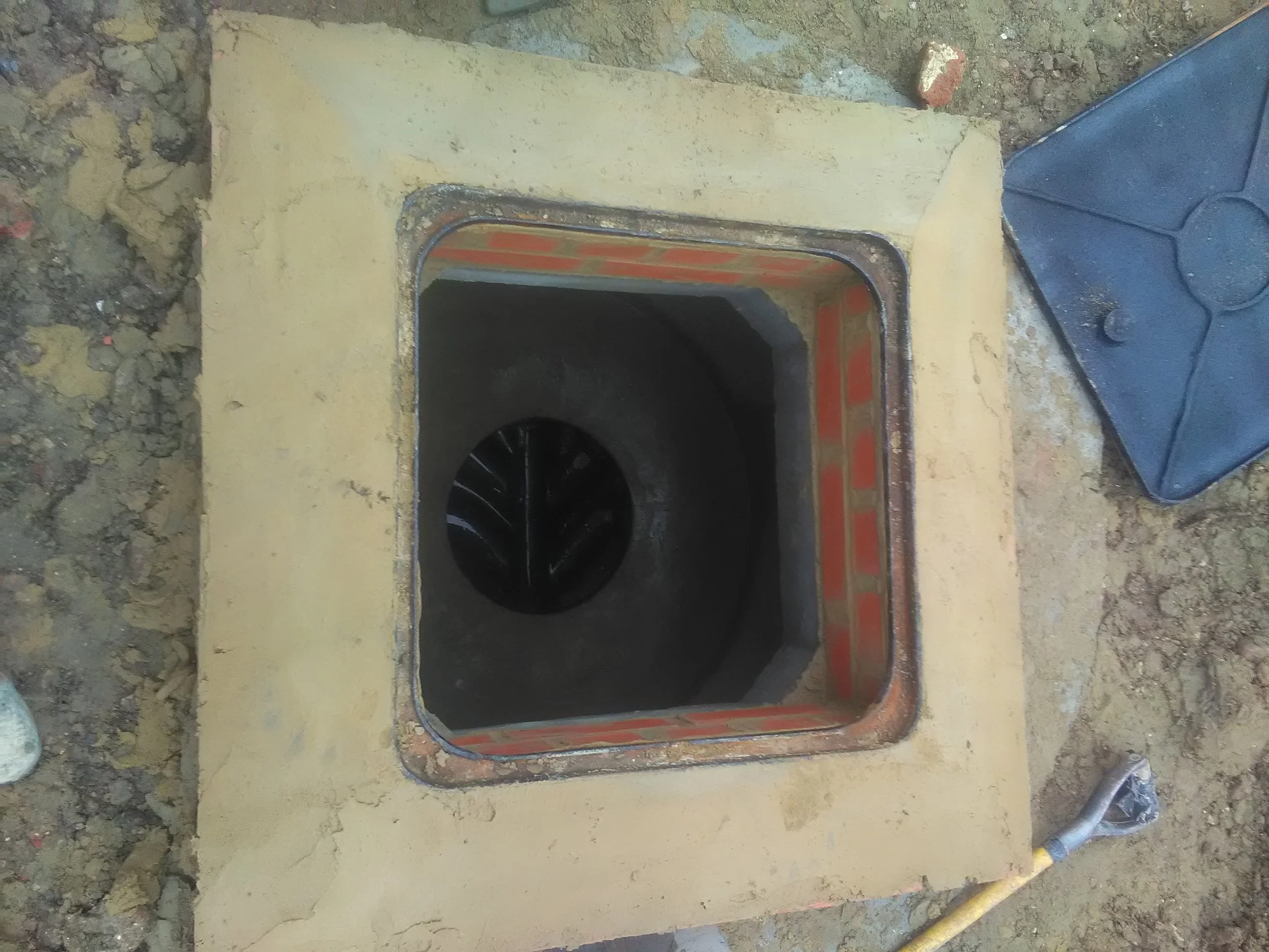 The image shows a rectangular hole in a concrete or cement surface, with one side surrounded by a yellowish material and some visible red bricks on the side. There are rough dirt and debris around the area, and a black drain or pipe is visible at the bottom inside the hole.