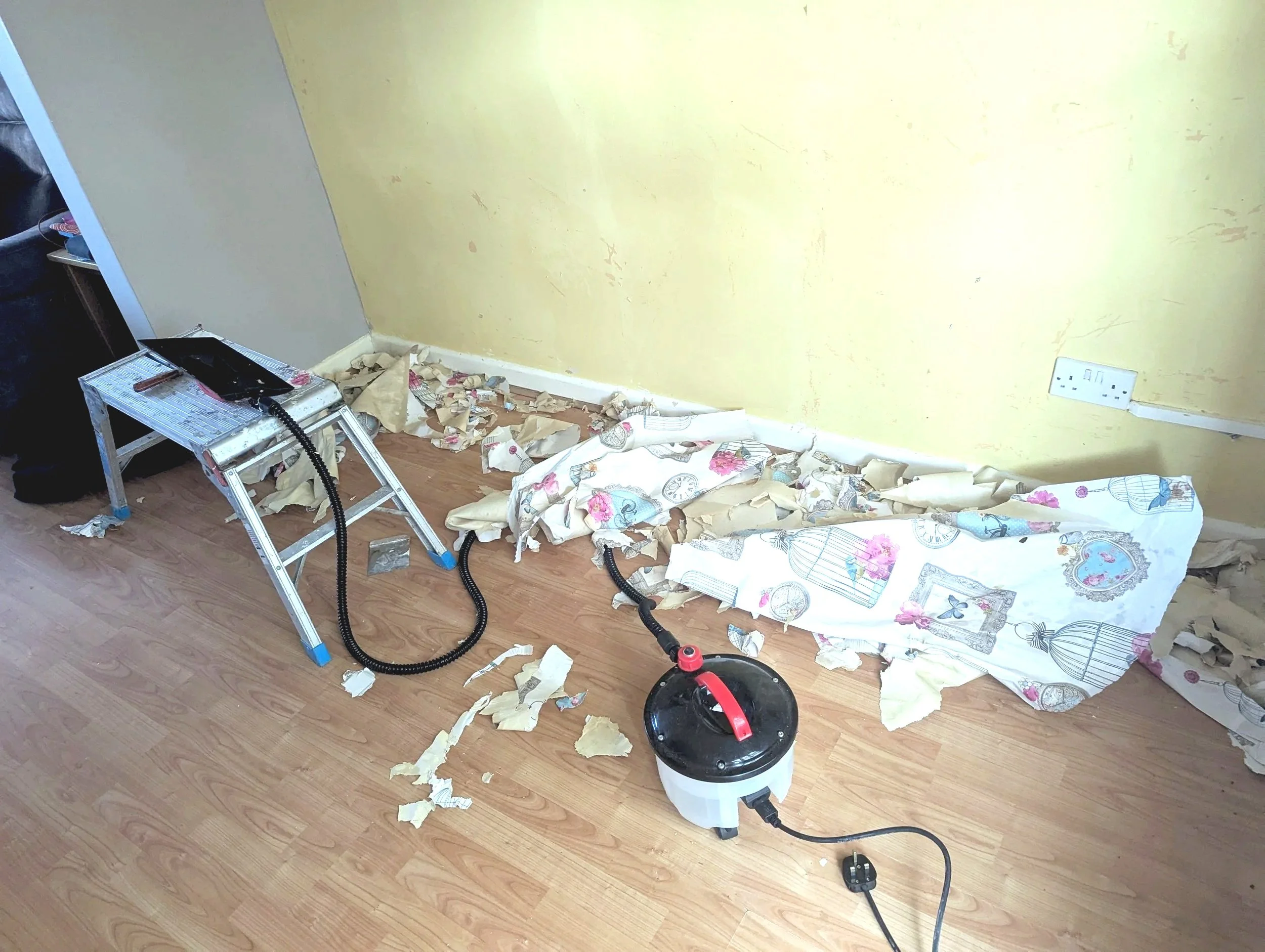 A partially renovated room with a yellow wall, a patch of gray wall, and scattered debris on the wooden floor, including torn wallpaper, a small step ladder, a vacuum cleaner, and some other items.