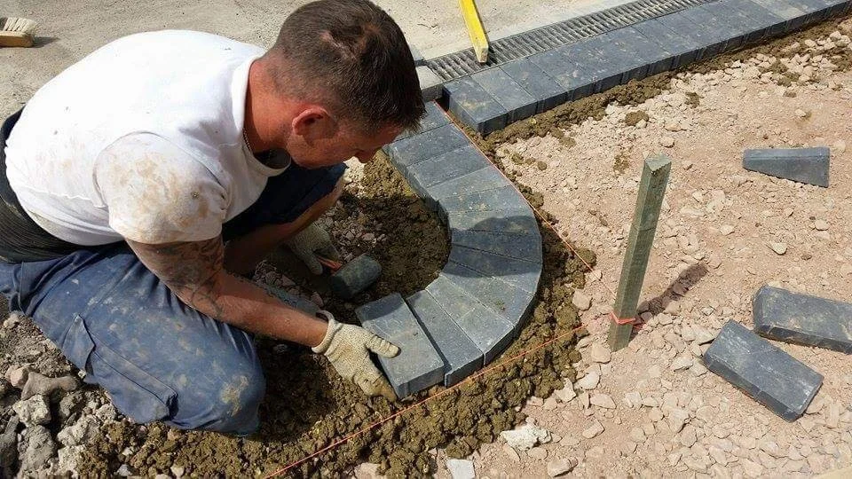 A man building a curved stone pathway or border by laying rectangular gray stones on a bed of dirt, with a string line for alignment.