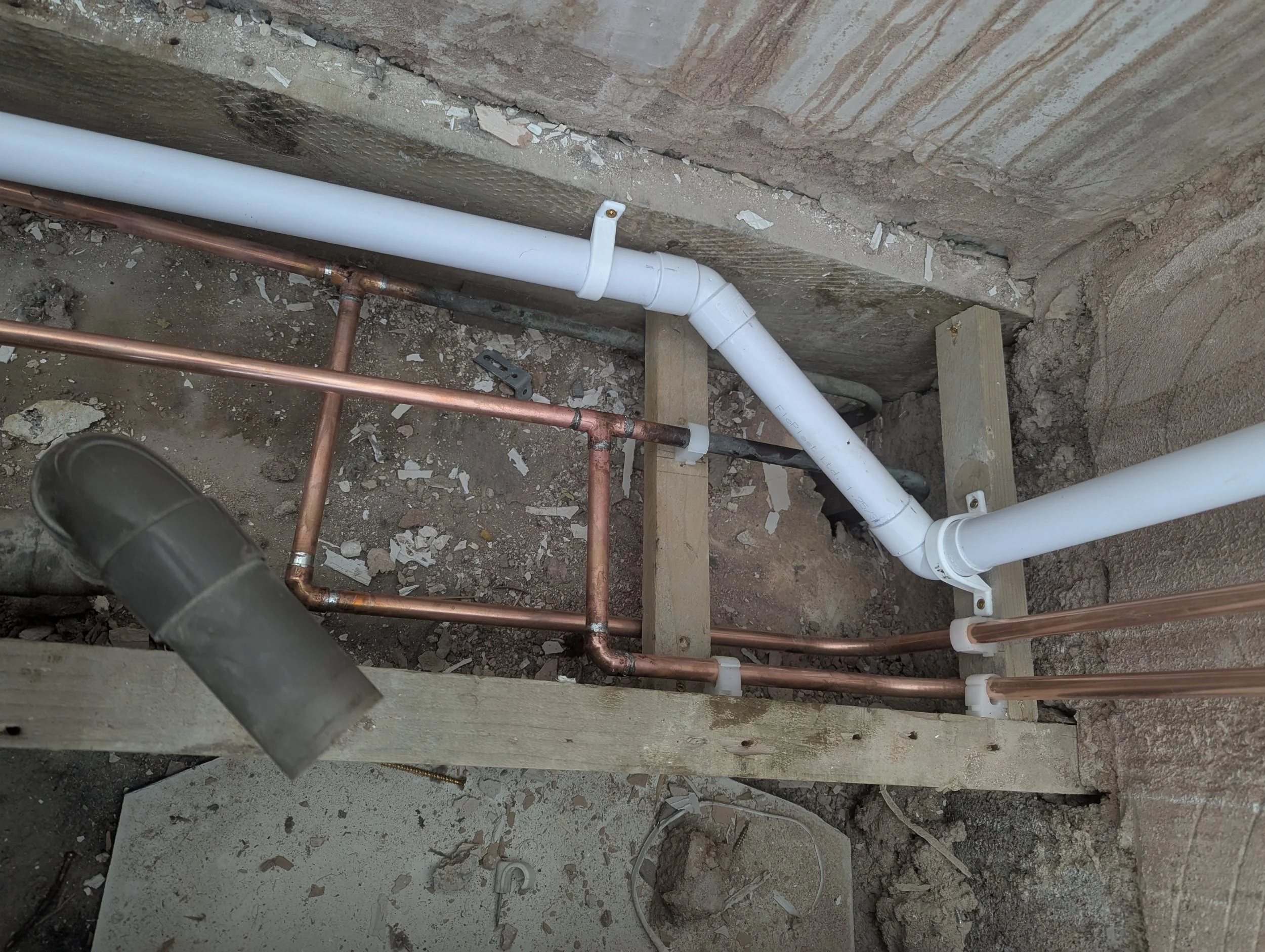 Pipes, including white PVC and copper, installed along a concrete wall in a construction area, with dirt and debris on the ground.