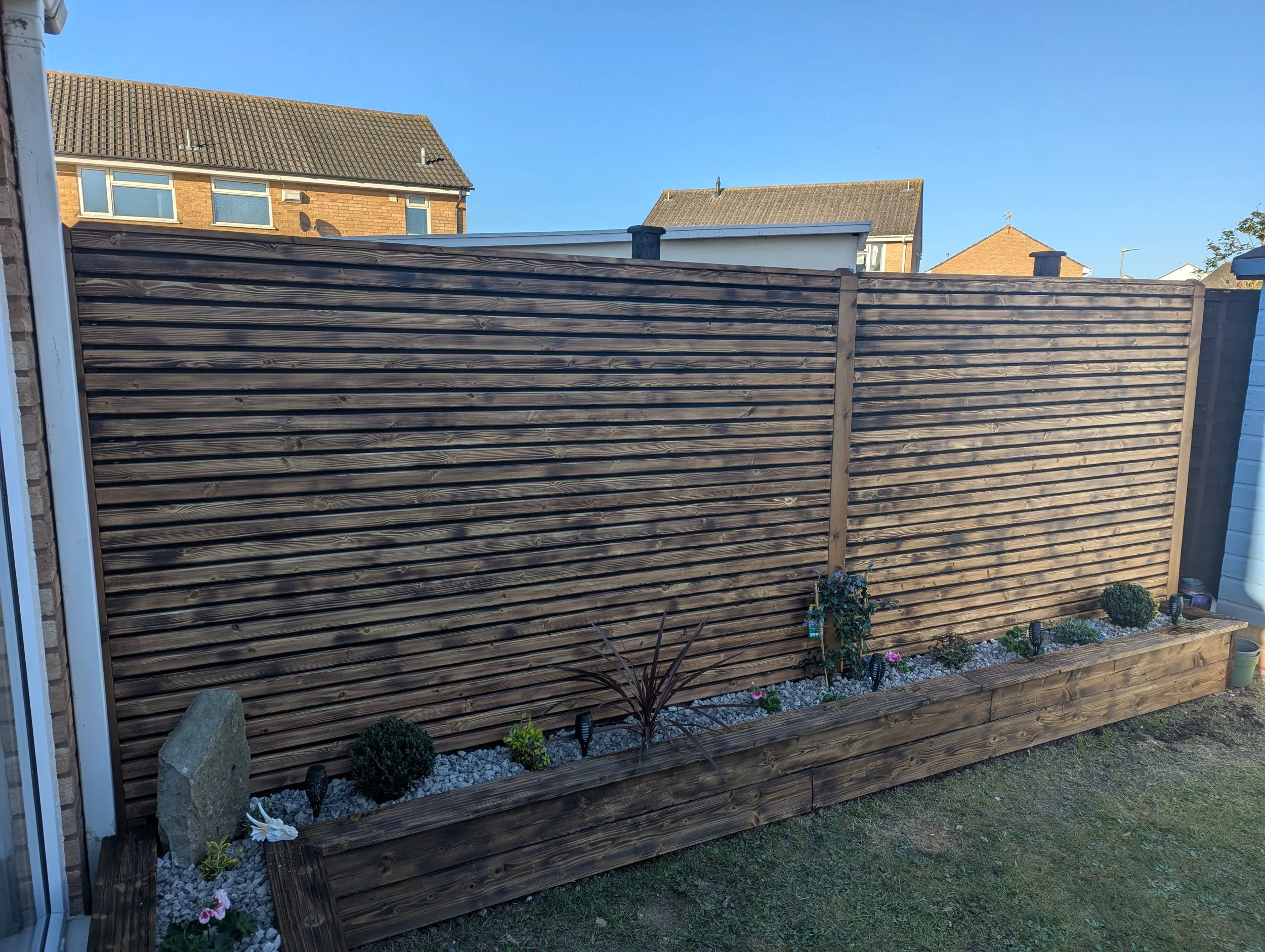 A wooden privacy fence with a flower bed at the base, containing small plants and flowers, in a residential back garden