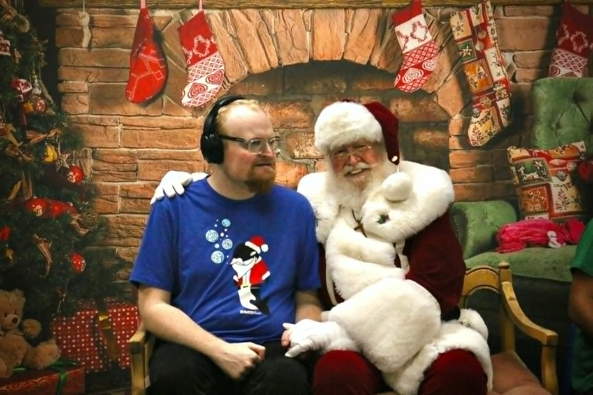 A man with glasses wearing a blue Christmas sweater and a bearded Santa Claus in a red suit and white beard sitting together in front of a decorated brick fireplace with Christmas stockings hanging. They are indoors, celebrating Christmas.