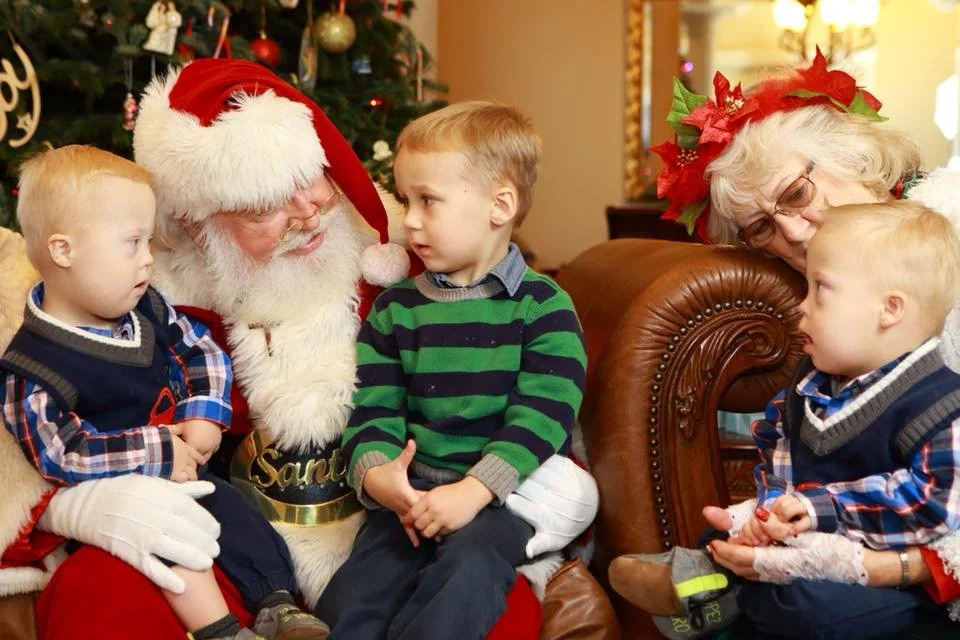 A Santa Claus with children during Christmas, sitting next to a woman with poinsettia headband, inside a decorated room with a Christmas tree.