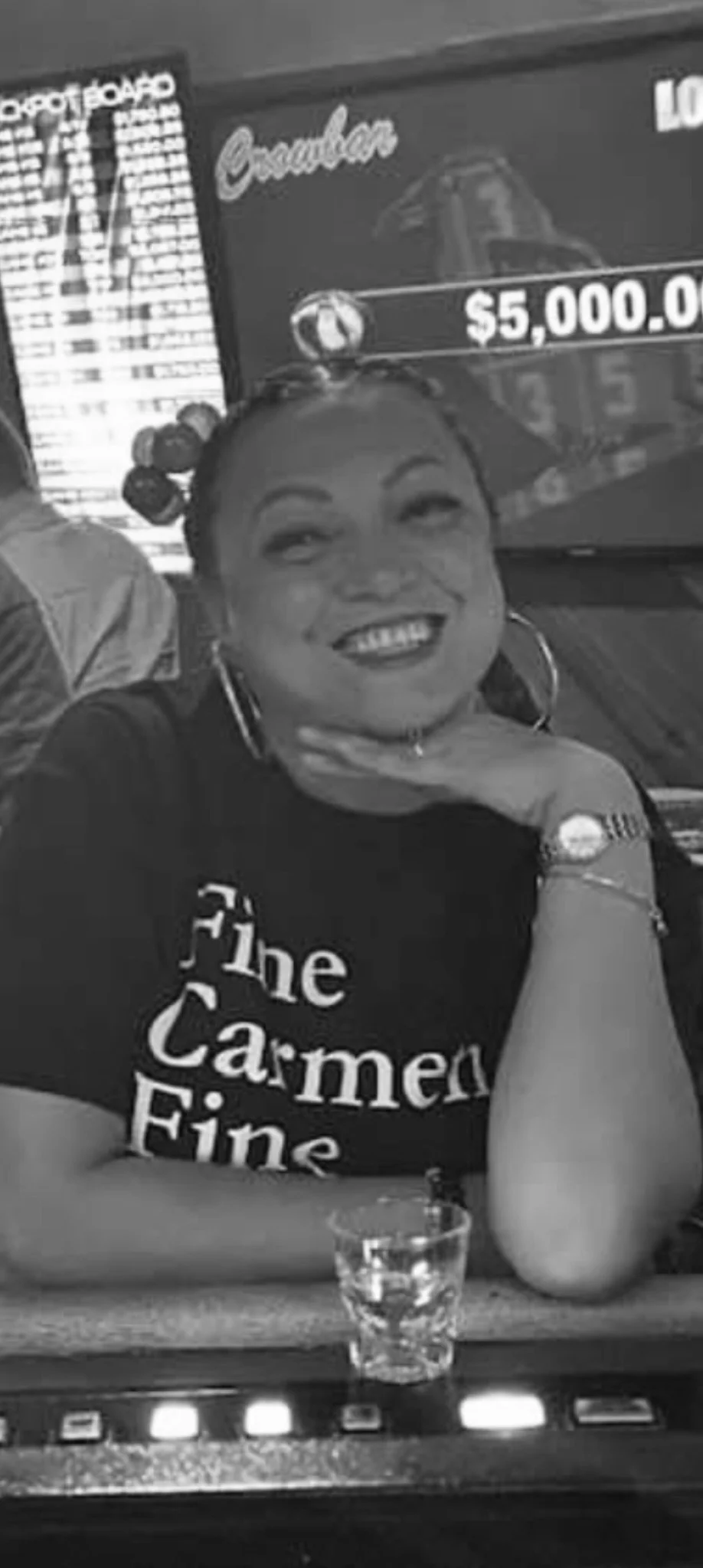 A smiling woman with curly hair styled in buns, wearing hoop earrings, and a black T-shirt with white text that reads "The Carmela Fine". She is sitting at a table with a shot glass in front of her, and her hand is resting on her chin. In the background, there is a digital display showing a gambling payout of $5,000, and a sign with the word "Barbados." The scene appears to be inside a casino or bar.