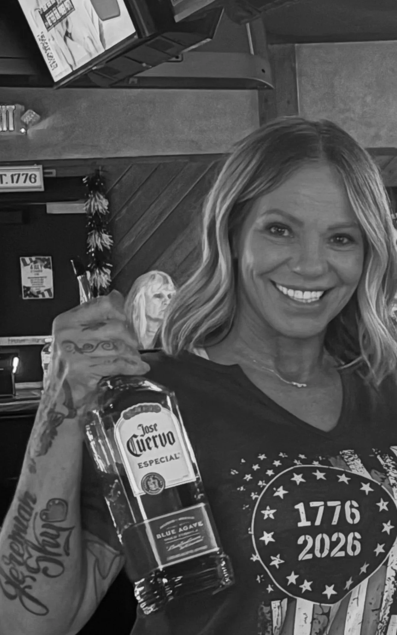 A woman with blonde hair smiling and holding a bottle of Jose Cuervo tequila, with tattoos on her arm. There is another woman in the background, and the setting appears to be a bar or restaurant.