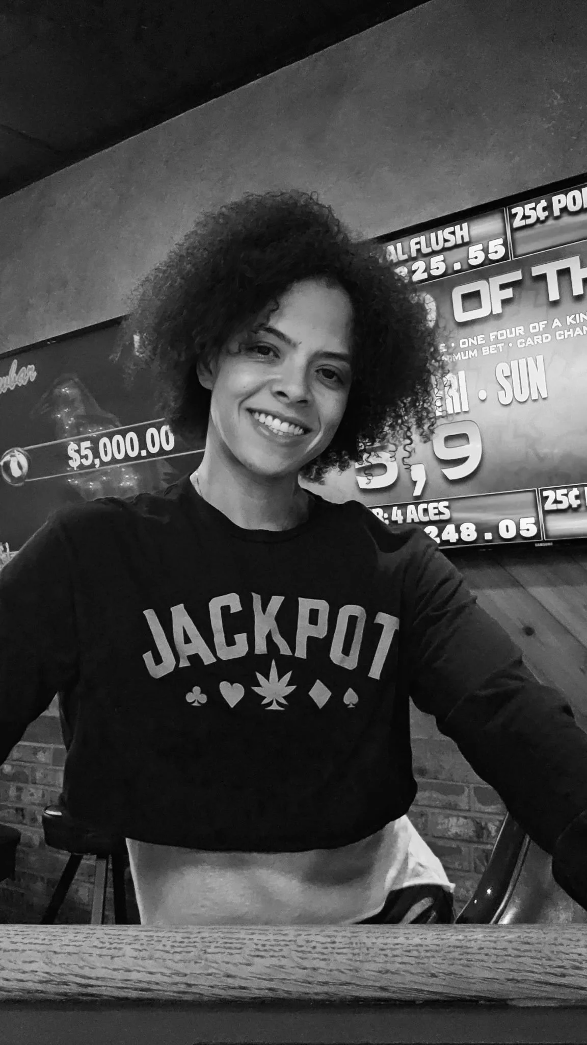 A woman with curly hair smiling, wearing a black shirt with the word 'JACKPOT' and playing card symbols, sitting at a table in a restaurant or bar with a brick wall and a gaming sign in the background.