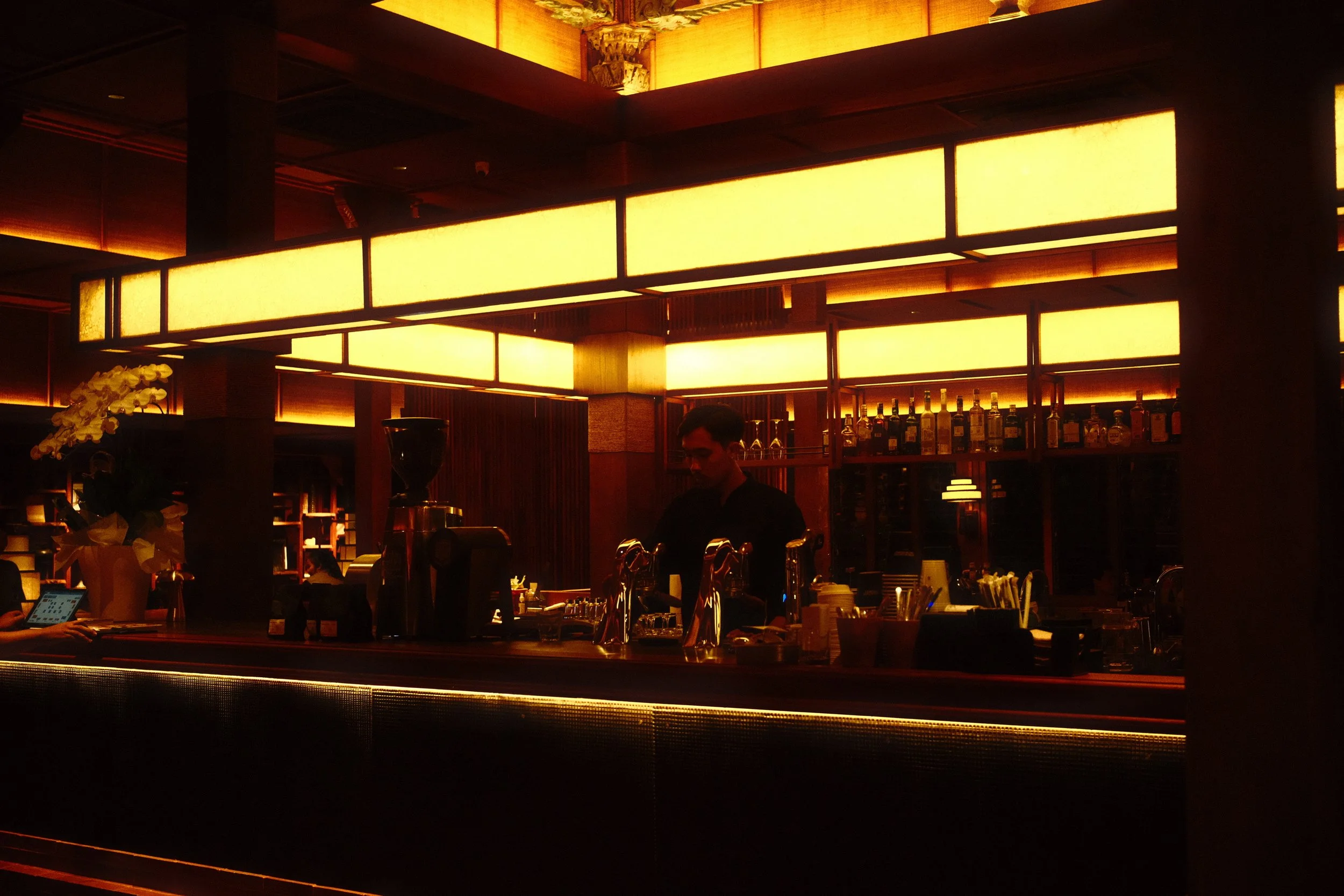 An interior of a dimly lit restaurant or bar with warm yellow lighting fixtures and a bar counter with a bartender and bar tools, alongside decorative elements like flowers.