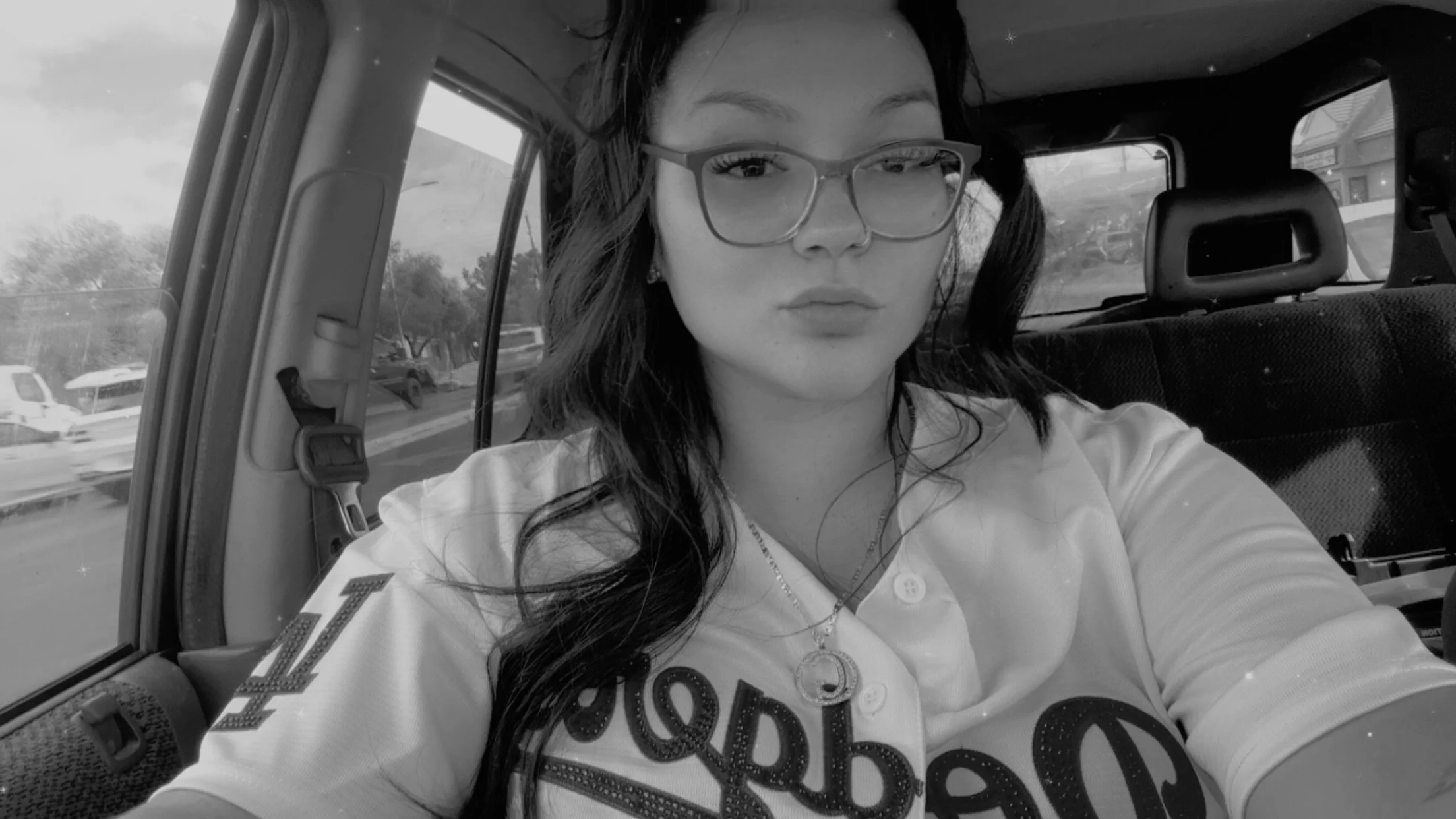 Black and white photo of a young woman with glasses, long dark hair, and wearing a shirt with printed text, sitting inside a vehicle with visible window and headrest, facing the camera.