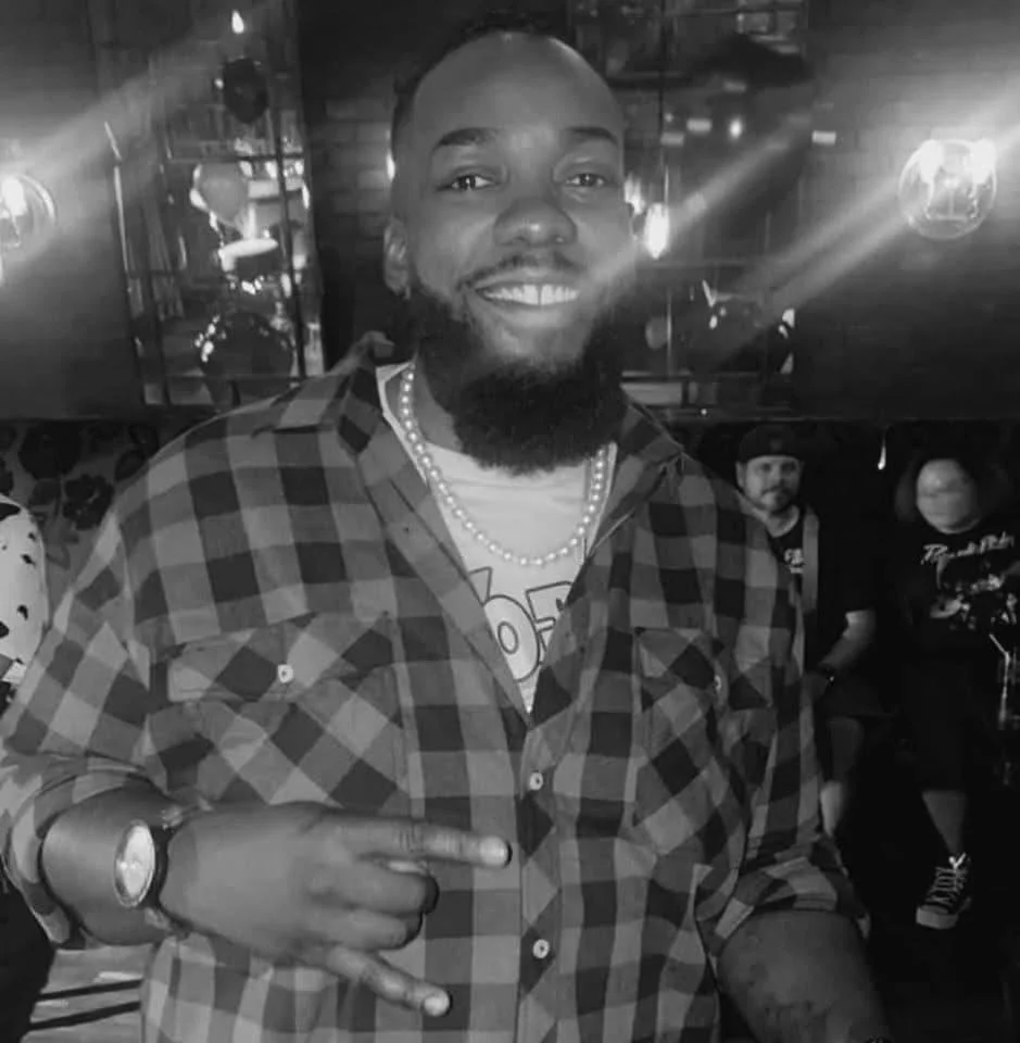 A smiling man with a beard wearing a checkered shirt, a pearl necklace, and a watch, pointing to himself in a lively, dimly lit bar or club. People and disco lights are visible in the background.