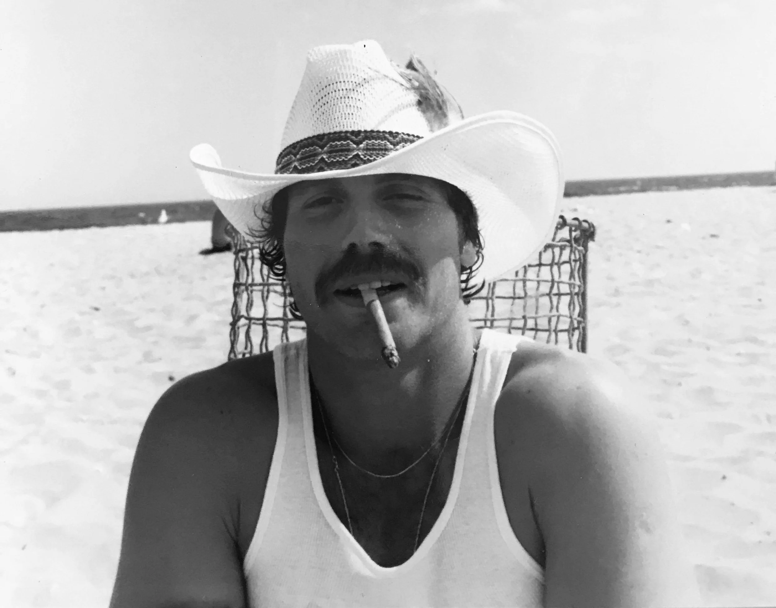 A man wearing a cowboy hat smoking a cigarette at the beach.