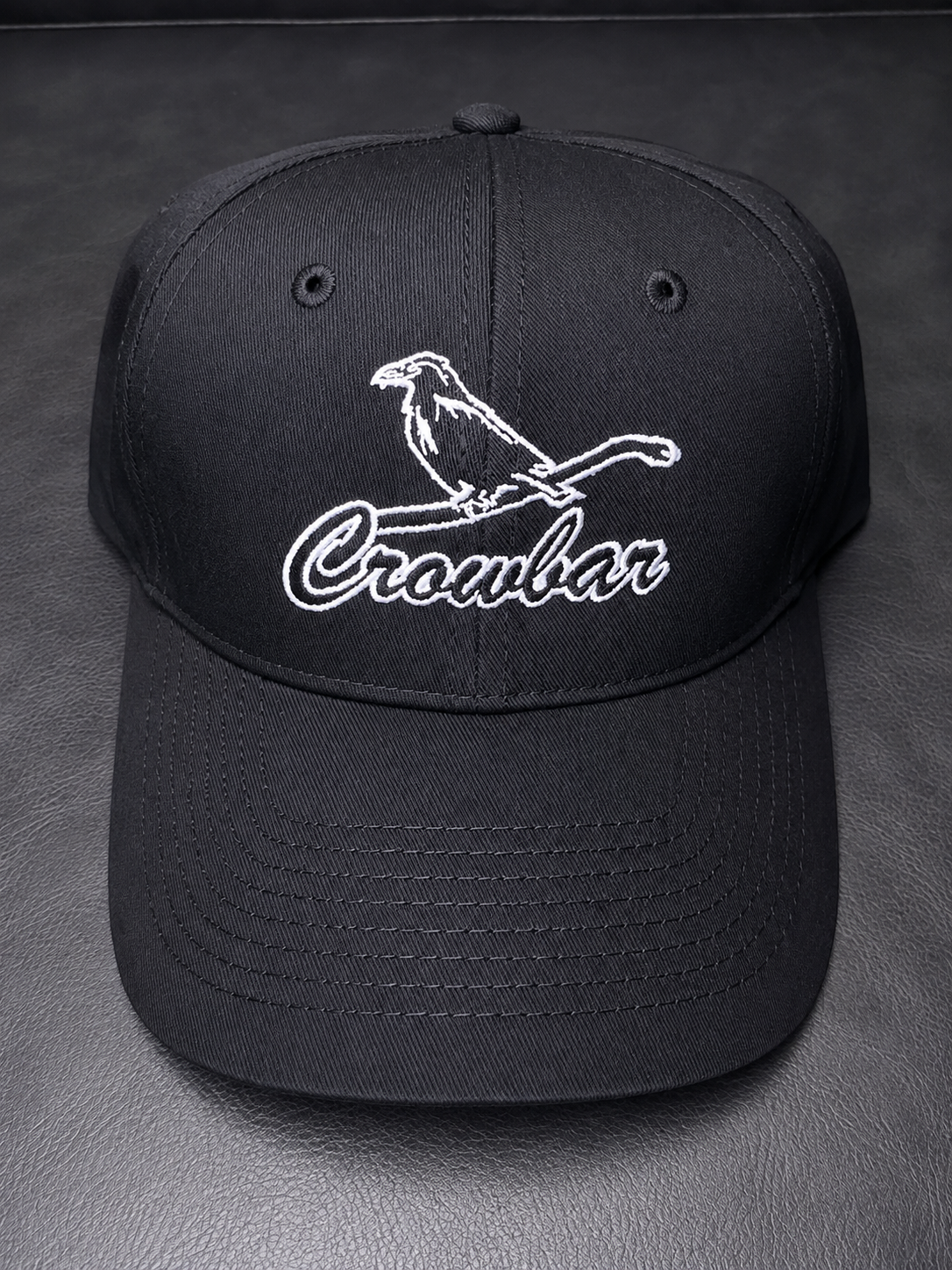 Black baseball cap with white embroidered logo of a bird perched on a branch and the word "Crocbar" in cursive.
