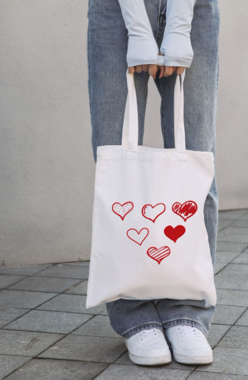 Person standing on a sidewalk holding a white tote bag with red and black heart designs.