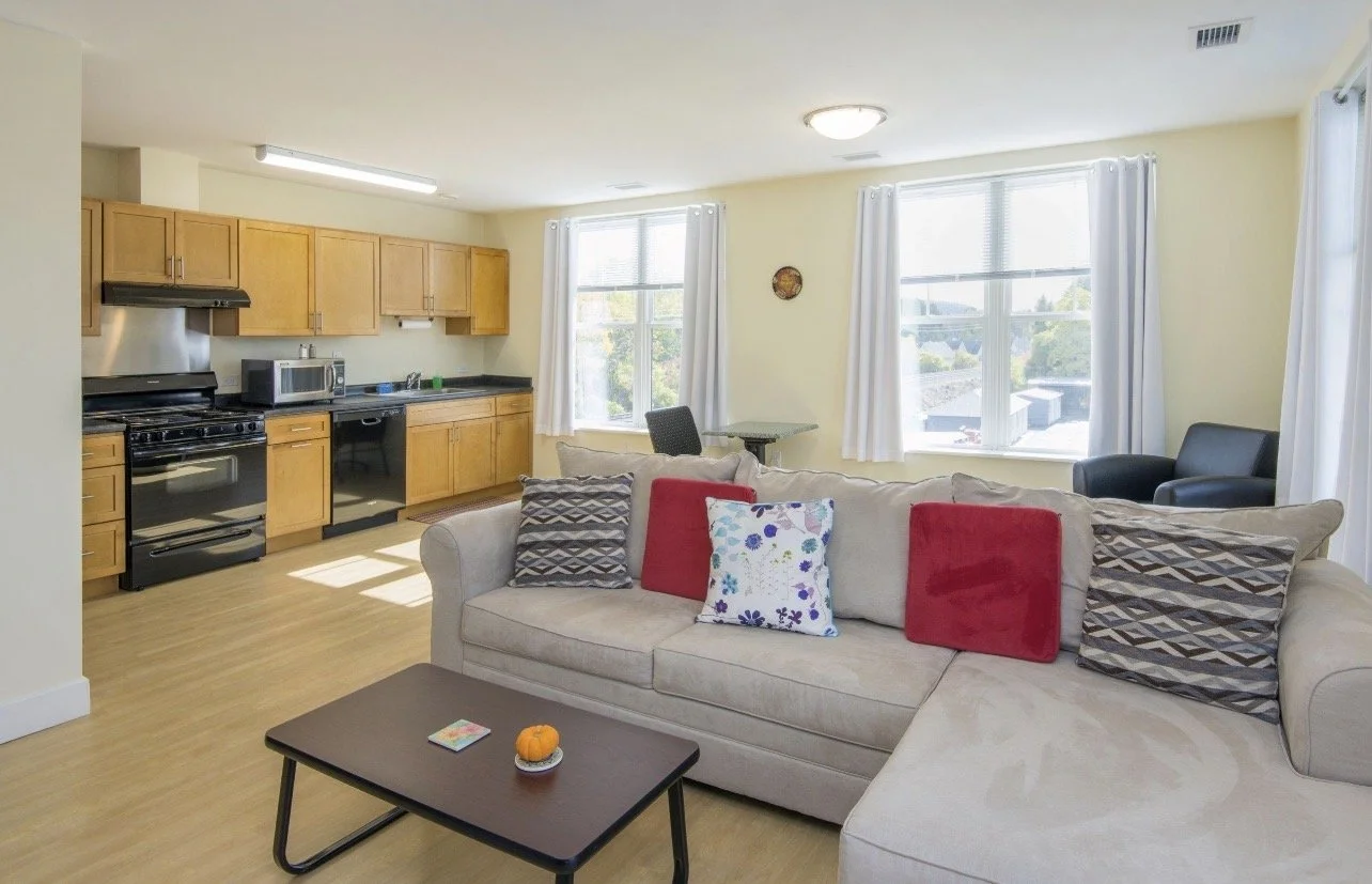 Living room with a beige couch, colorful pillows, a black coffee table with a small pumpkin, and large windows with white curtains. Behind the couch, there's an open kitchen with wooden cabinets, black appliances, and a microwave.
