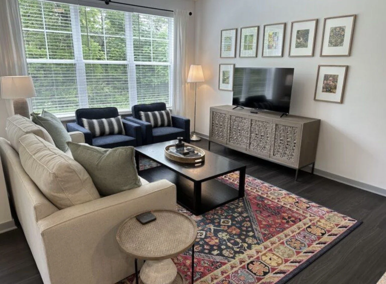 Living room with large window, white couch with pillows, two black armchairs with pillows, rectangular coffee table, patterned rug, TV on a decorative wooden stand, framed pictures on the wall, floor lamp, small side table with a remote can be seen.