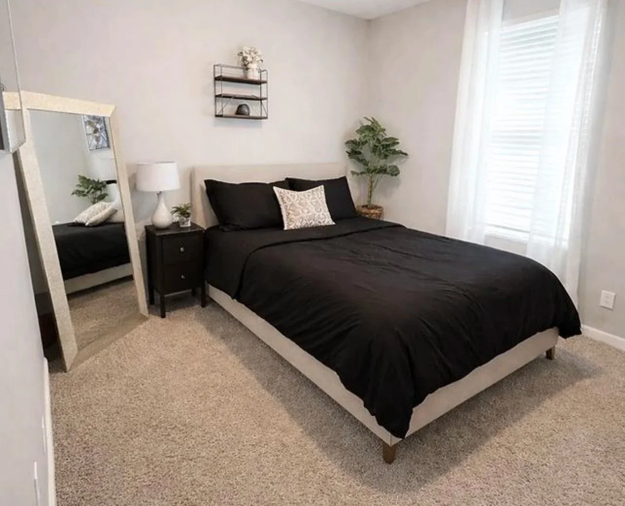 A bedroom with a bed covered in black bedding, a white headboard, two black pillows, and a decorative cushion. There is a small black nightstand with a white lamp and small plant, a full-length mirror leaning against the wall, a potted plant in the corner, and sheer white curtains over a window.