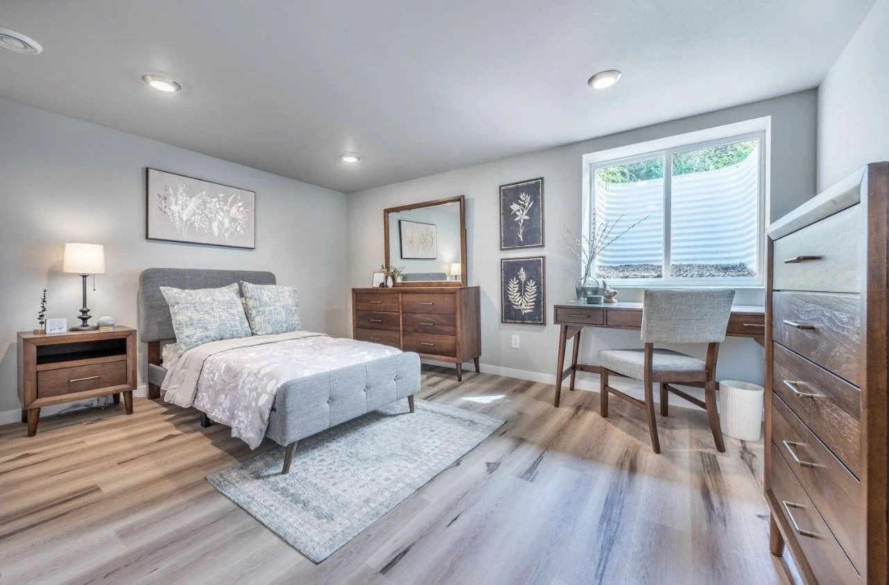 A bedroom with a bed, nightstand, dresser, mirror, and a desk with a chair. The room has hardwood floors, light gray walls, and a window with blinds.