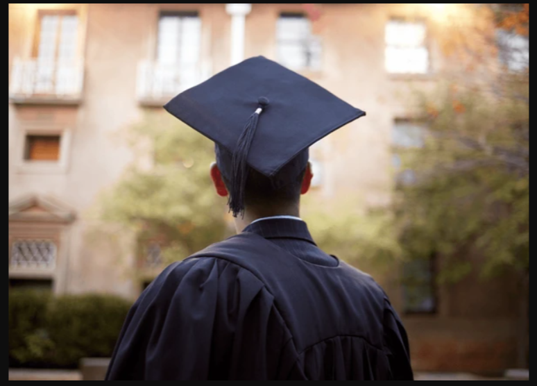 A graduate in a cap and gown facing away, outdoors on a college campus