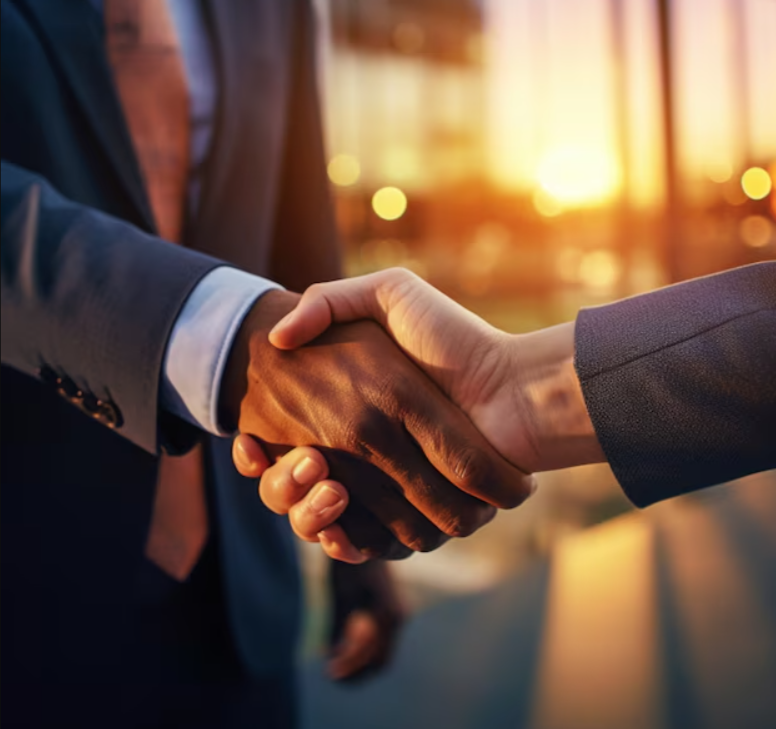 Two people in business suits shaking hands during sunset in a modern office or conference room.