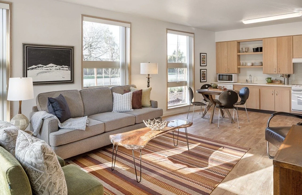 Living room with sofa, wooden coffee table, armchair, patterned area rug, kitchen with cabinets, dining table, and chairs, and windows allowing natural light.