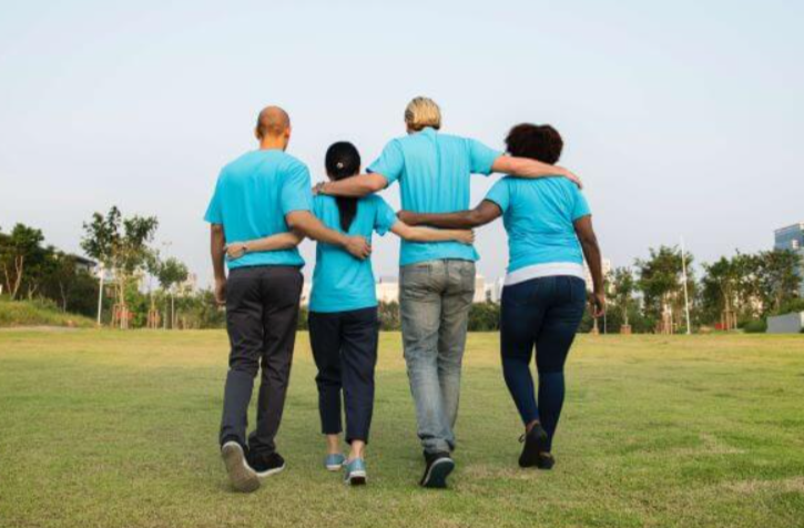 Four diverse people in matching blue shirts walking together arm-in-arm in a park.