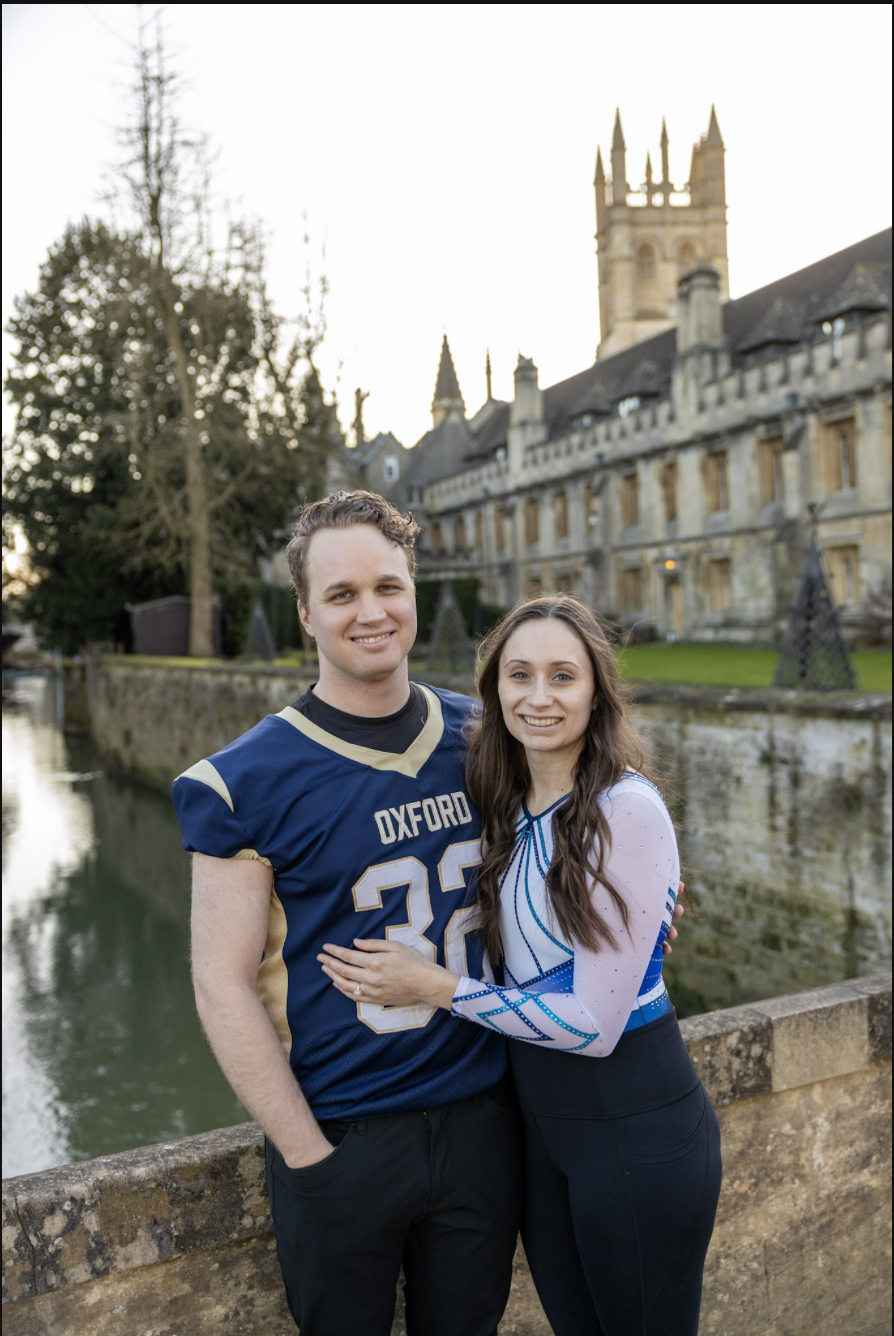 A smiling couple stands near a stone bridge over a canal in front of a historic castle or university building with towers and spires, in Oxford, England, during daytime.