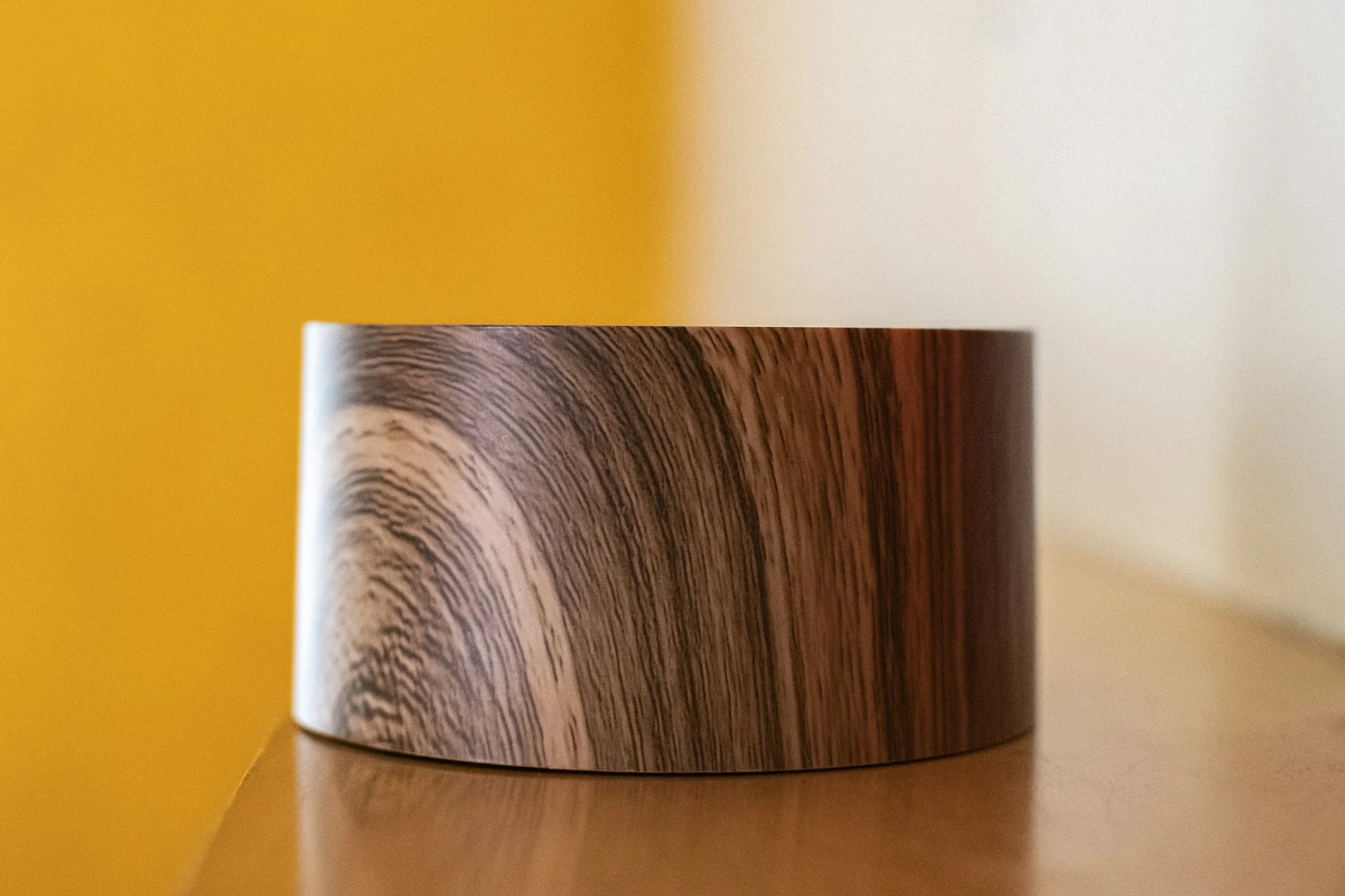 A close-up of a cylindrical wooden container with a wood grain pattern, placed on a wooden surface against a yellow background.