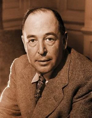 A man with a receding hairline, wearing a brown suit and tie, sitting indoors with a neutral expression.