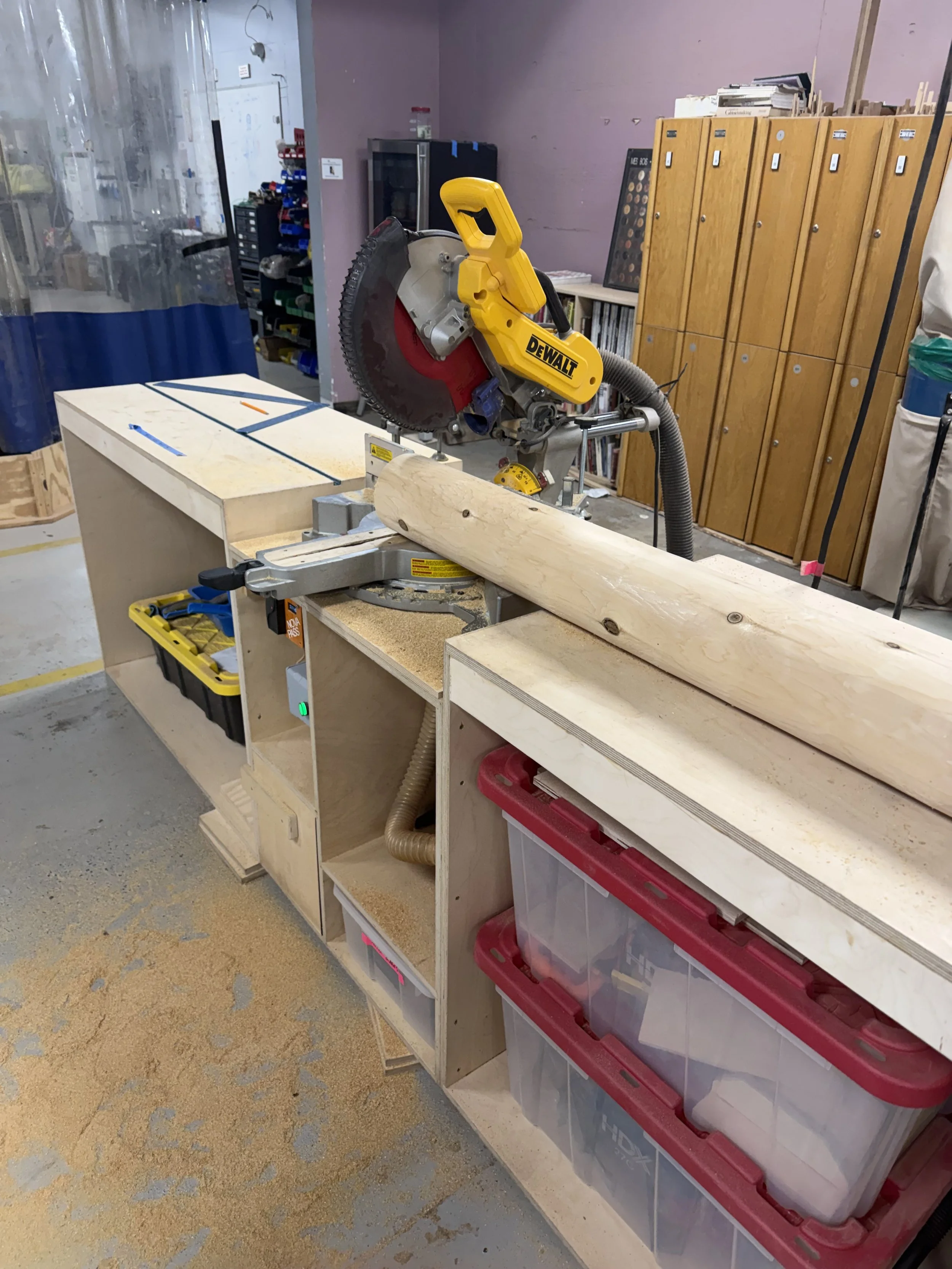 A workshop with a yellow DeWalt miter saw on a wooden workbench cutting a piece of wood. Shelving with tools and supplies in the background.