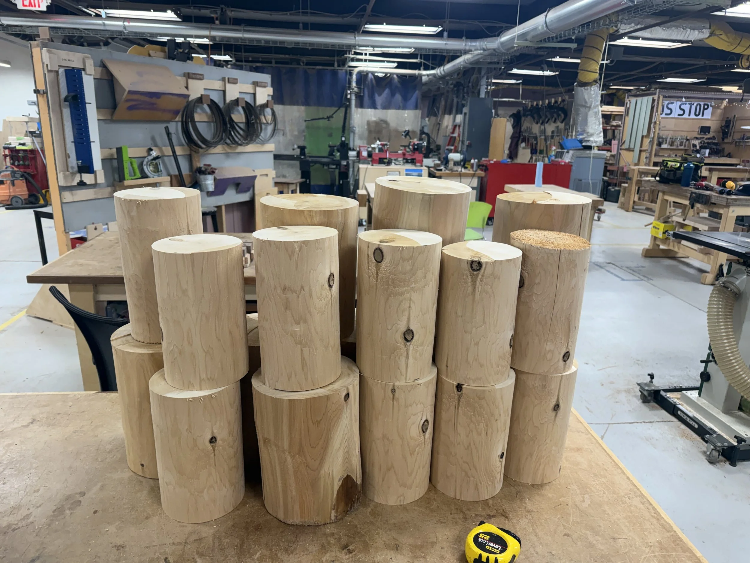 Stacked cylindrical wooden logs in a woodworking shop, with various tools and equipment visible in the background.